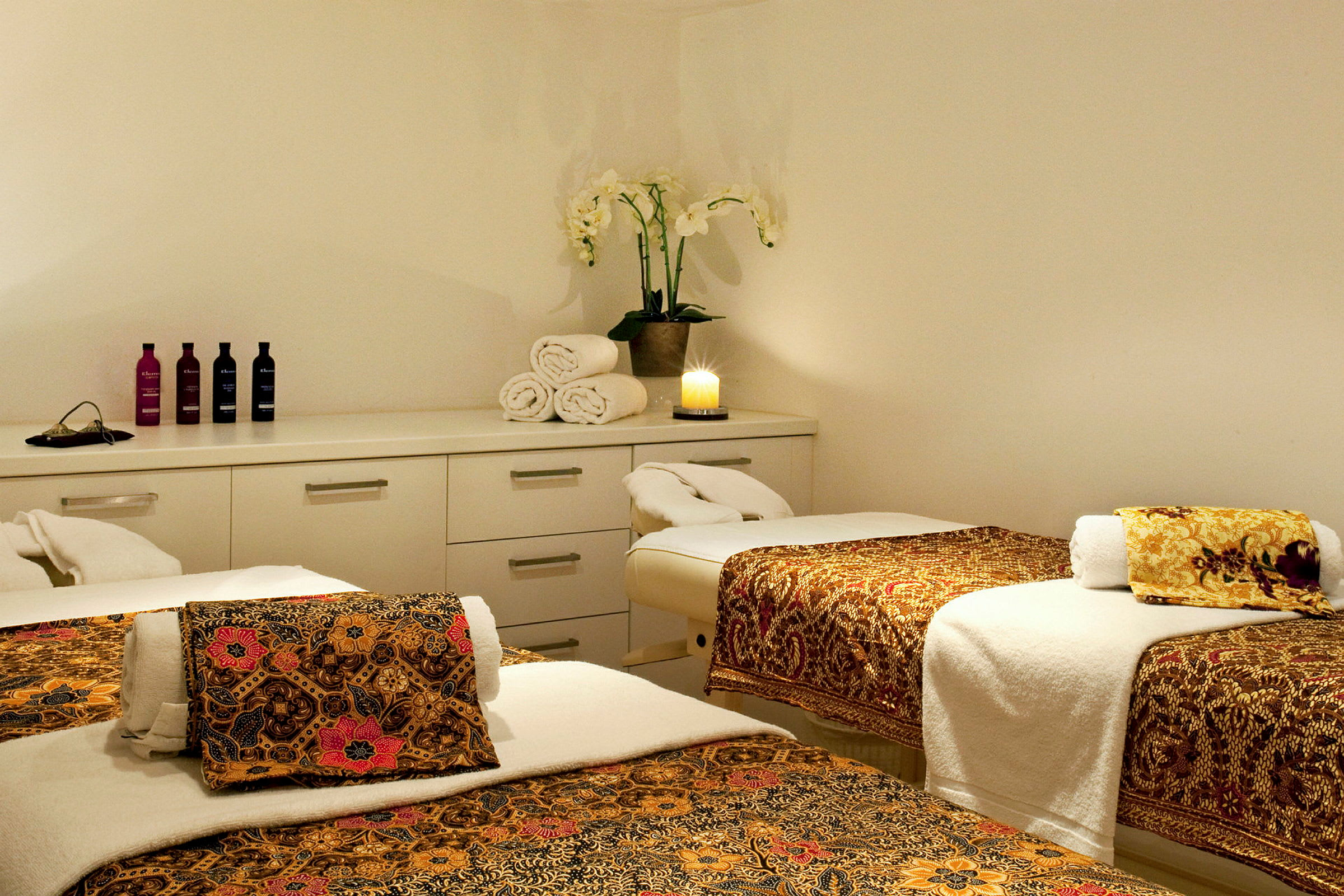Et spa-rum med tre massageborde dækket med stof med orientalske mønstre, håndklæder og en plante samt et tændt lys på en kommode. (AI-genereret tekst)