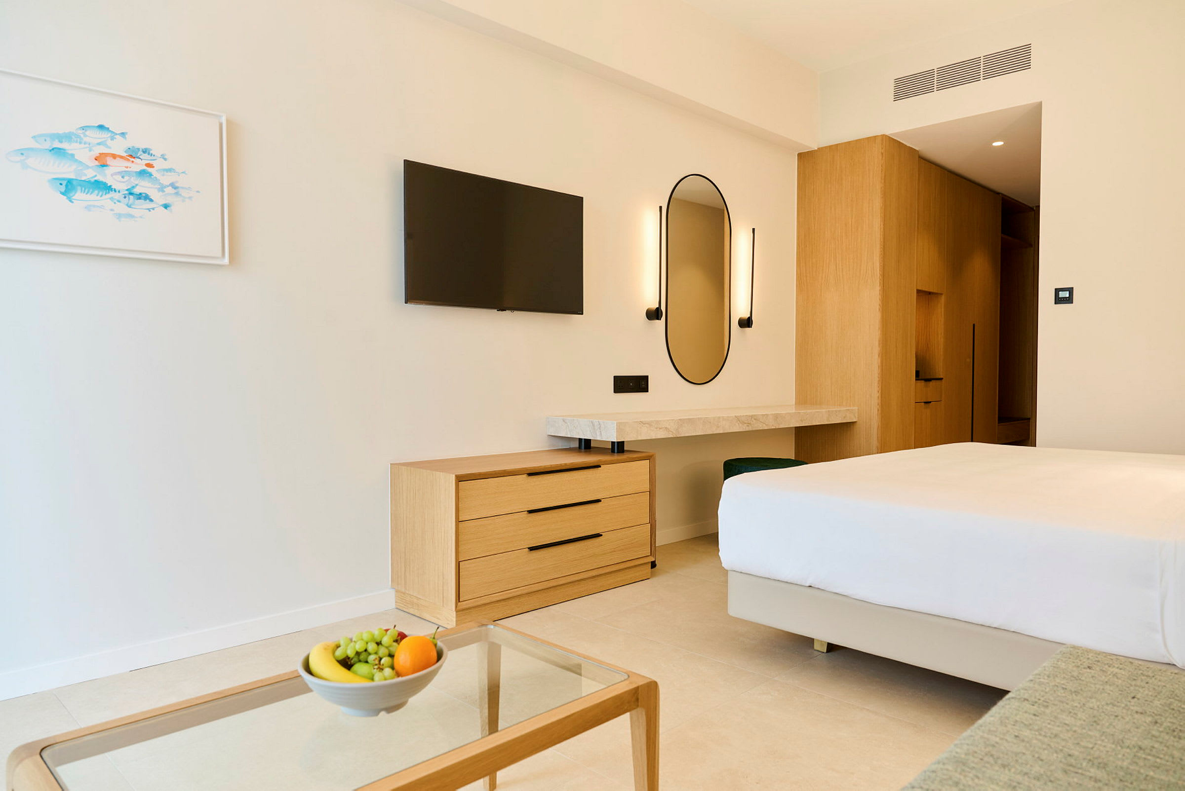 Et moderne hotelværelse med en dobbeltseng, en kommode, et vægmonteret TV og et skrivebord med spejl. På et glasbord står en skål med frugt. (AI-genereret tekst)