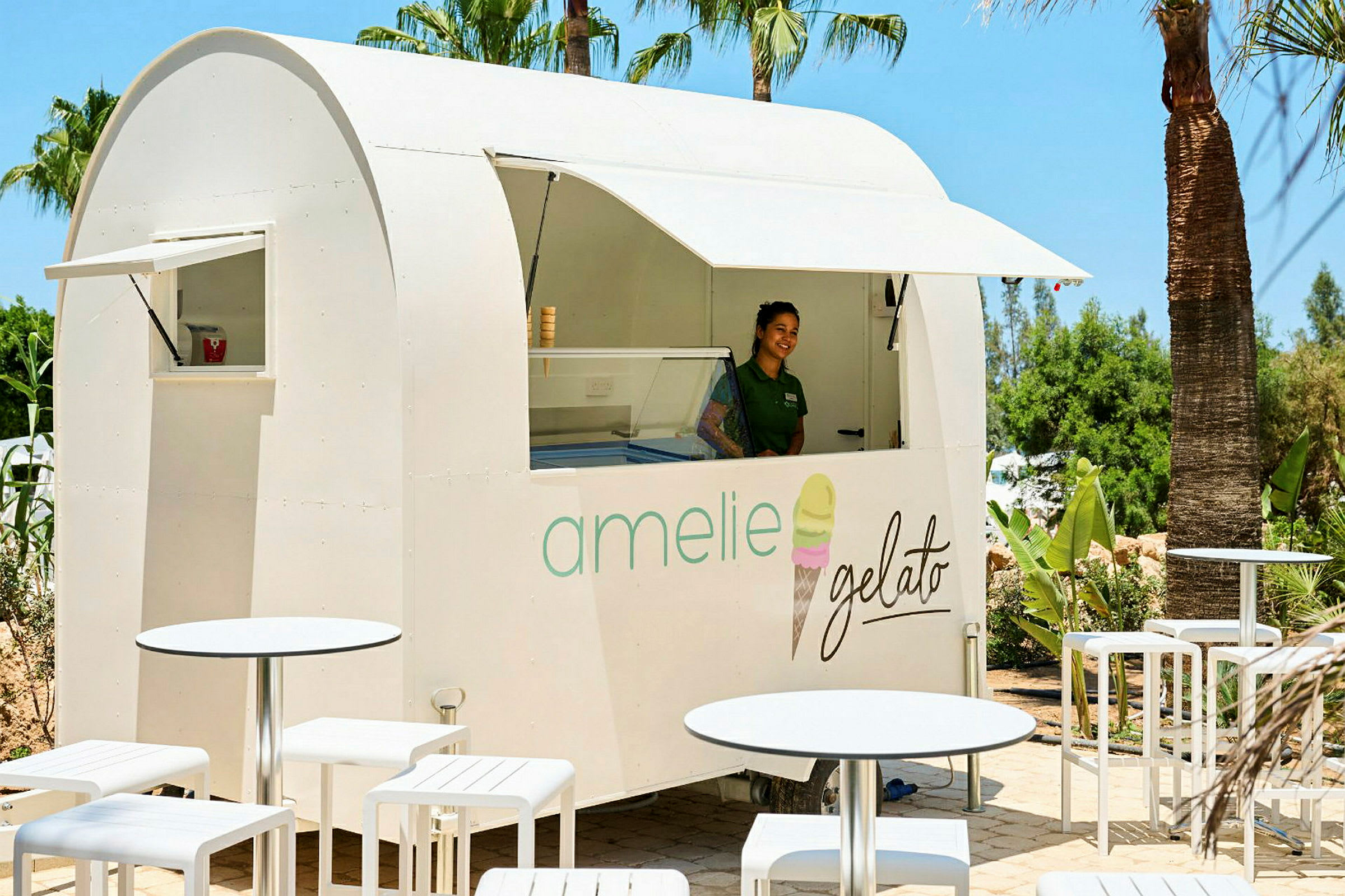 En hvid isbod med teksten "amelie gelato" står blandt palmer, omgivet af hvide havemøbler. (AI-genereret tekst)