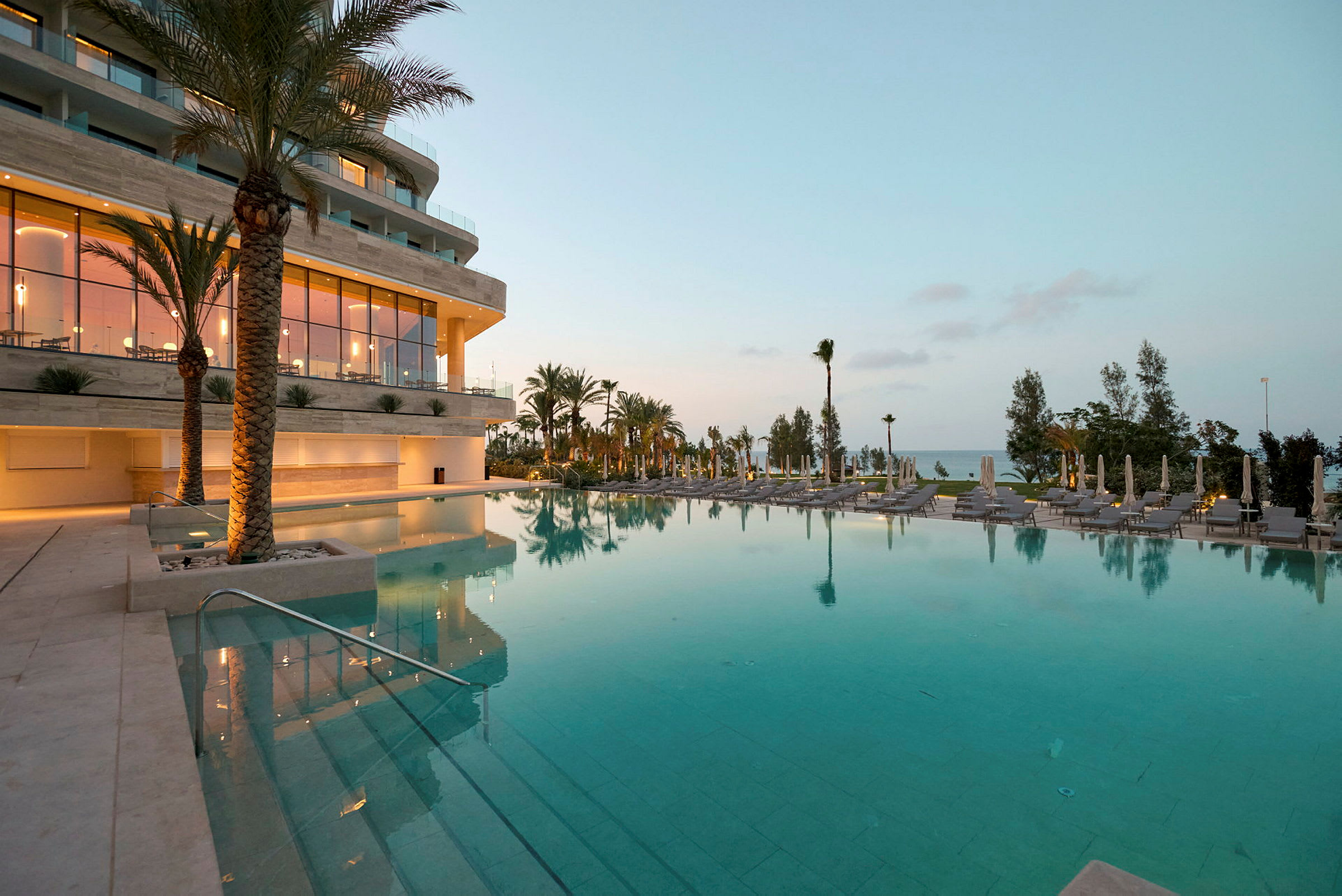 En stor udendørs pool omgivet af palmer ved siden af et moderne hotel om aftenen. (AI-genereret tekst)