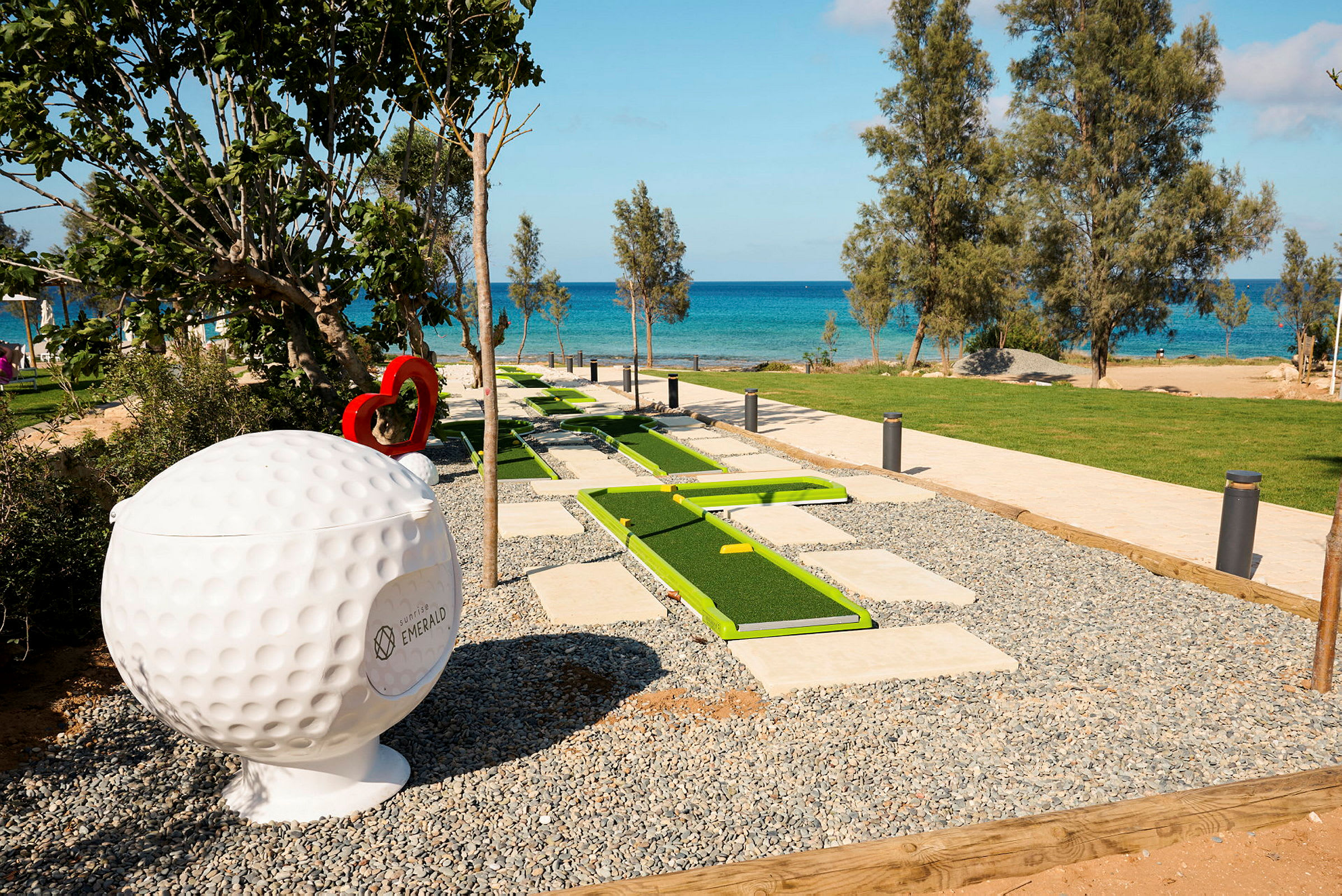 En minigolfbane med grønne baner er placeret nær stranden med blåt hav i baggrunden. En stor skulptur af en golfbold ses ved siden af. (AI-genereret tekst)