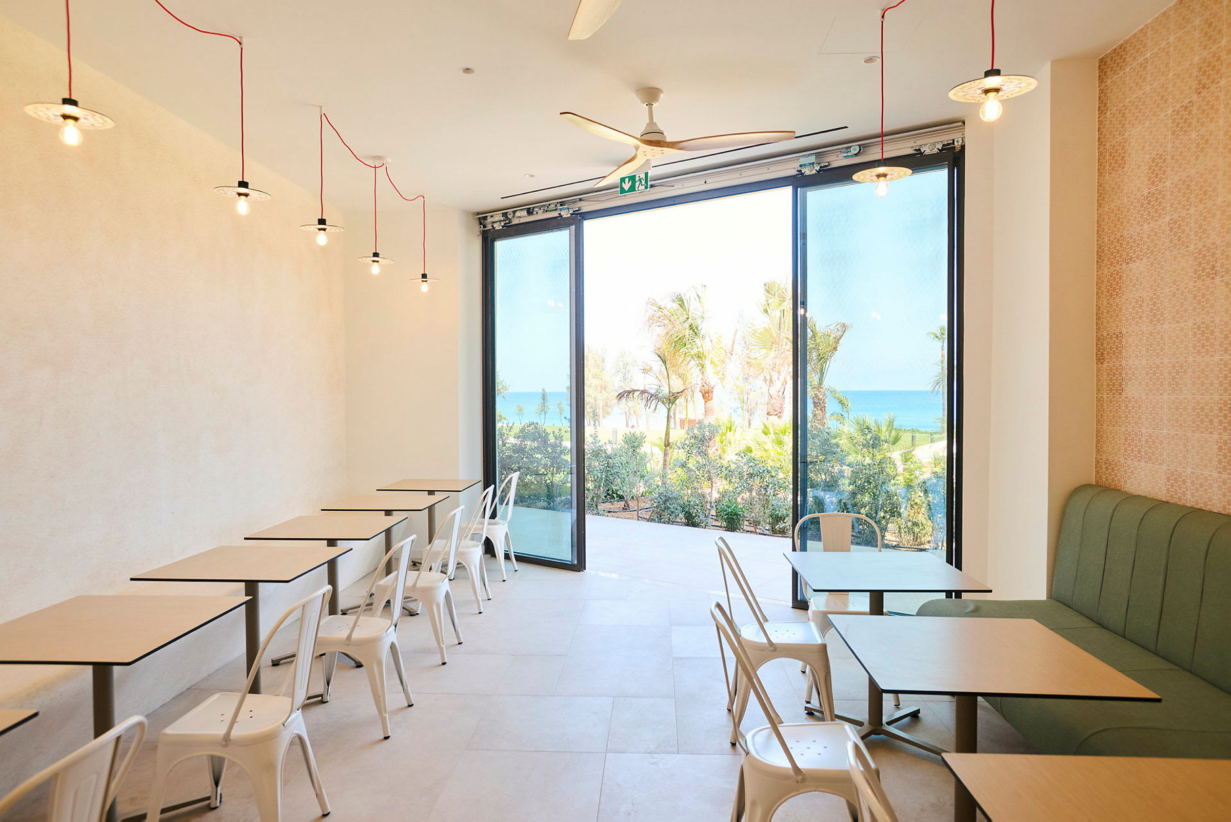 En caféinteriør med hvide stole og borde, store glasvinduer viser havet og grønne områder udenfor. (AI-genereret tekst)