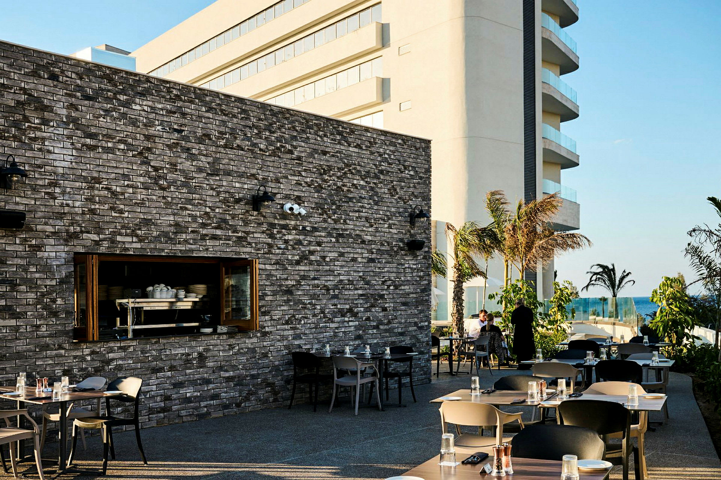 Udendørs terrasse ved en restaurant med borde og stole. En stor bygning ses i baggrunden, med palmer og havet i det fjerne. (AI-genereret tekst)