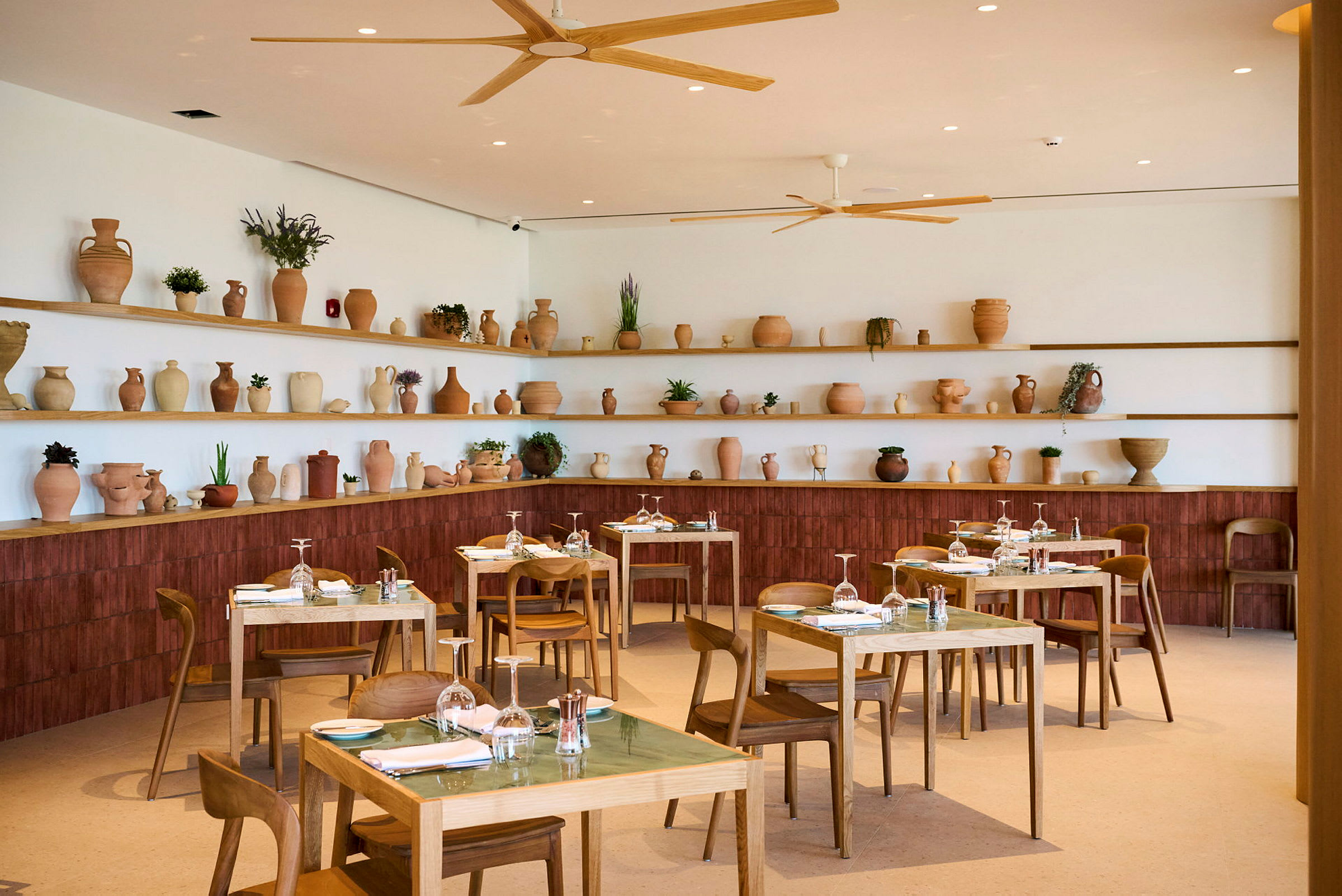 Billede af en restaurantindretning med træborde og stole. Hylder på væggen bagved er dekoreret med terrakottakrukker og planter. (AI-genereret tekst)