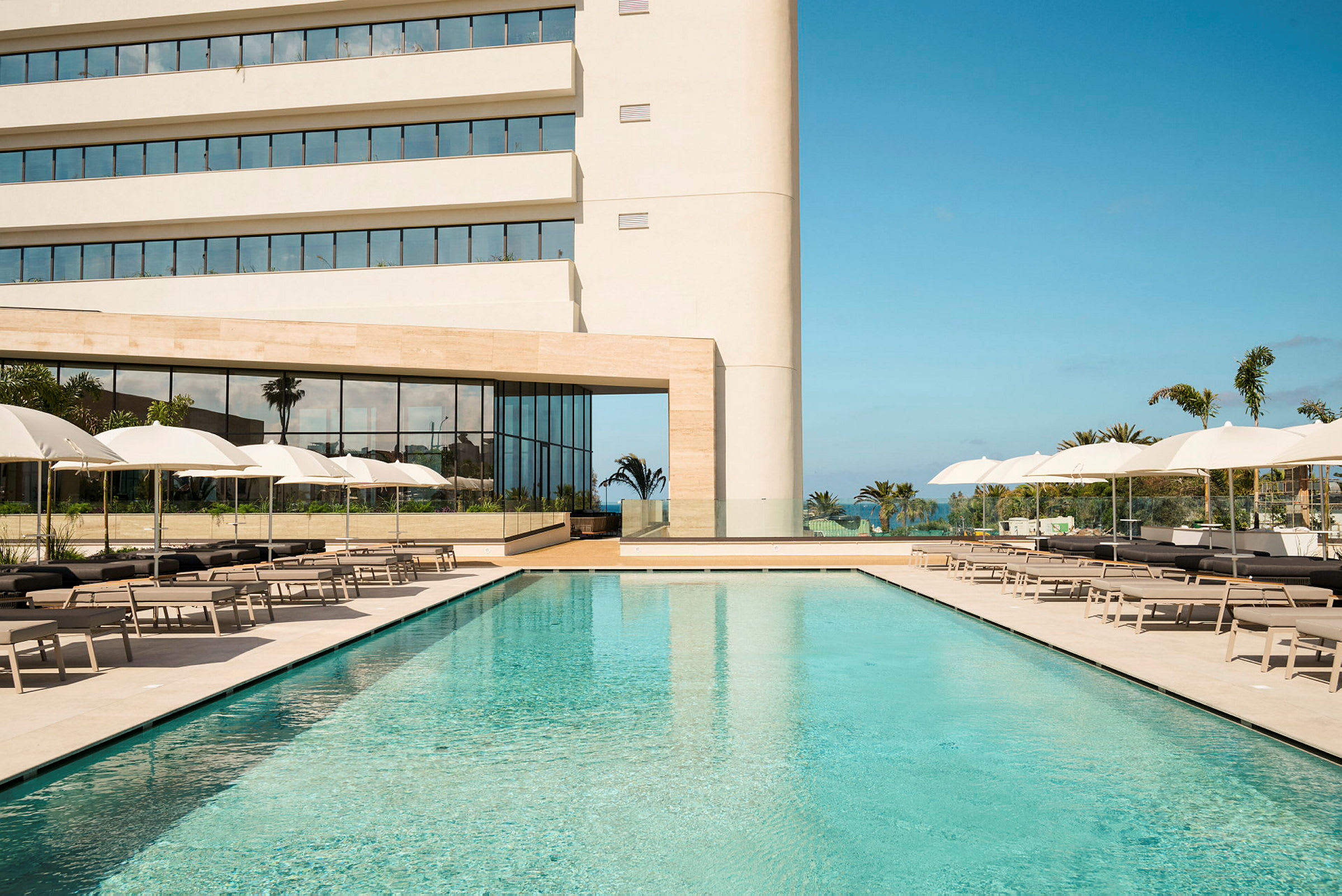 Hotel med pool, liggestole og parasoller foran en moderne bygning. (AI-genereret tekst)