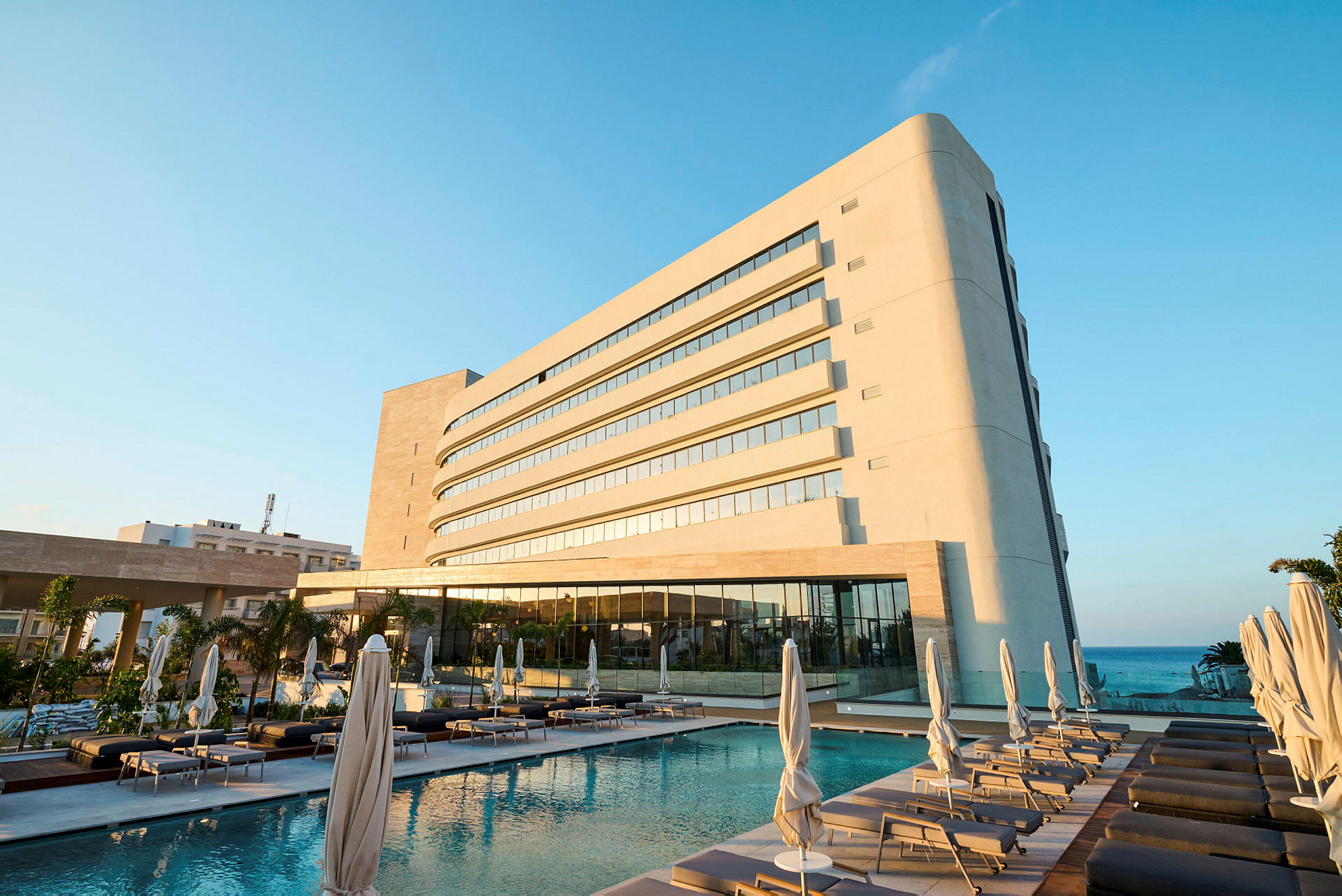En moderne hotelbygning ved siden af en pool med liggestole og parasoller. Havet kan ses i baggrunden. (AI-genereret tekst)