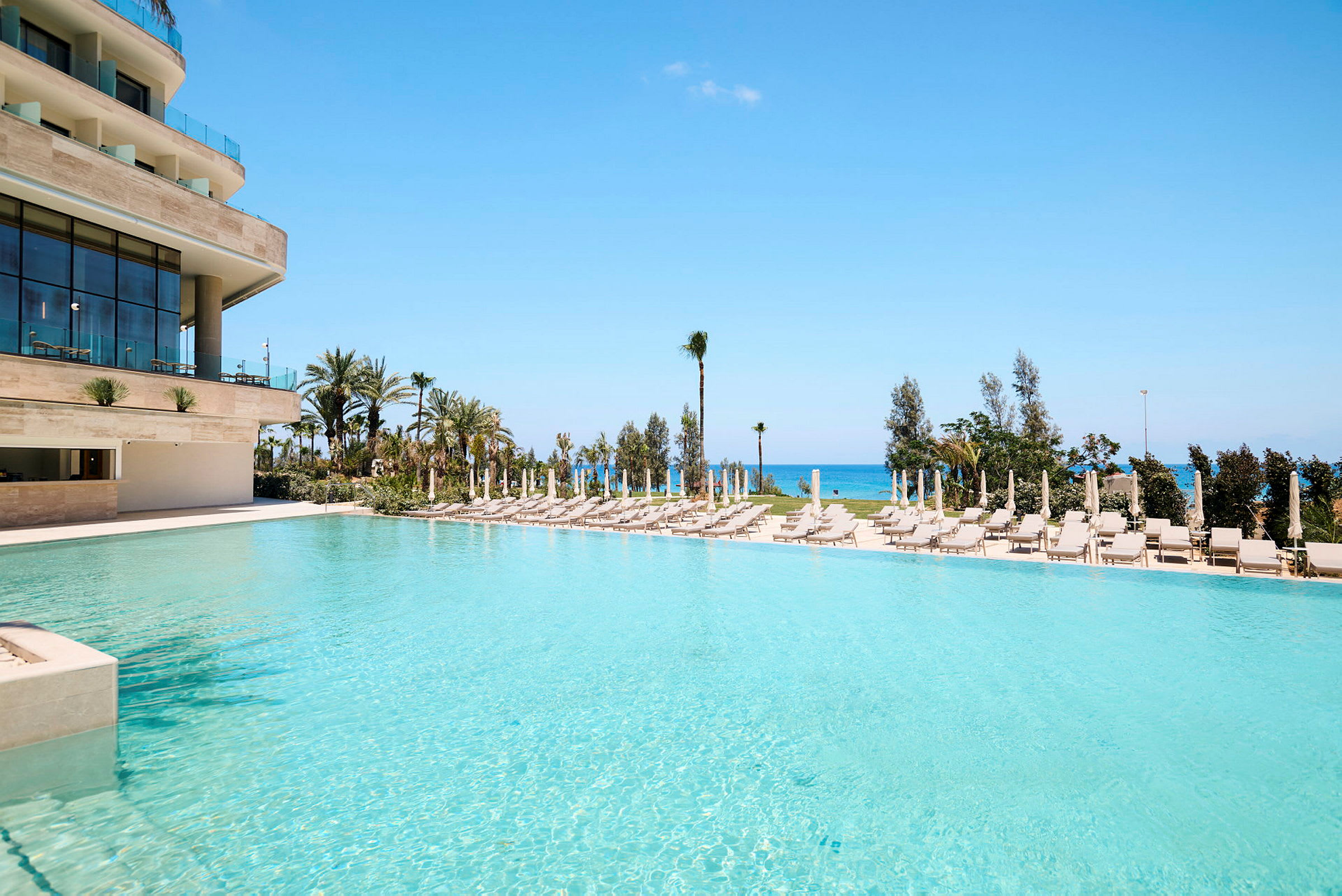 En swimmingpool ved en moderne bygning med solsenge og parasoller omkring, samt havet i baggrunden. (AI-genereret tekst)