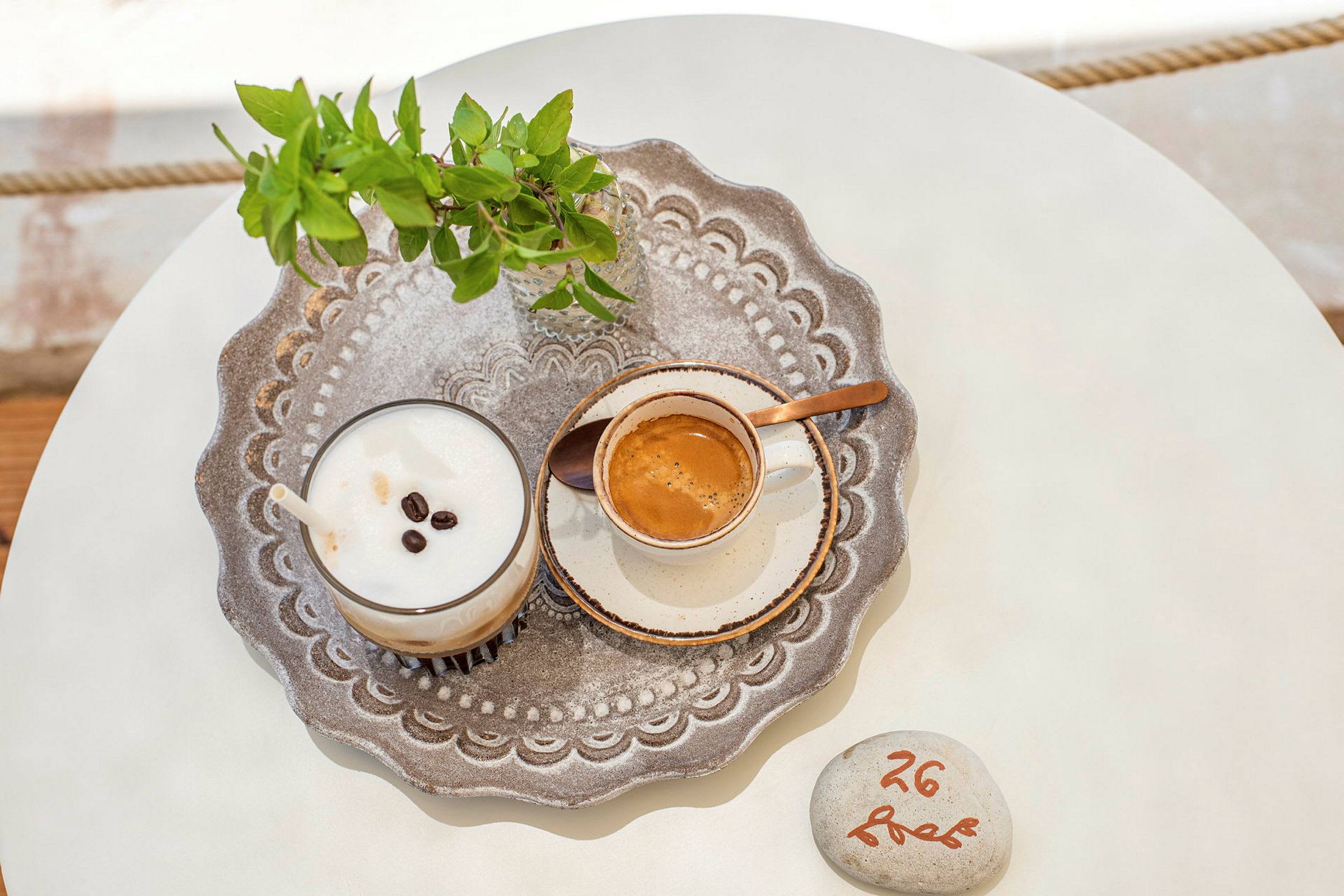 Et nærbillede af en bakke med en kop kaffe, en milkshake med kaffe og en lille glasvase med grønne blade på et bord. (AI-genereret tekst)