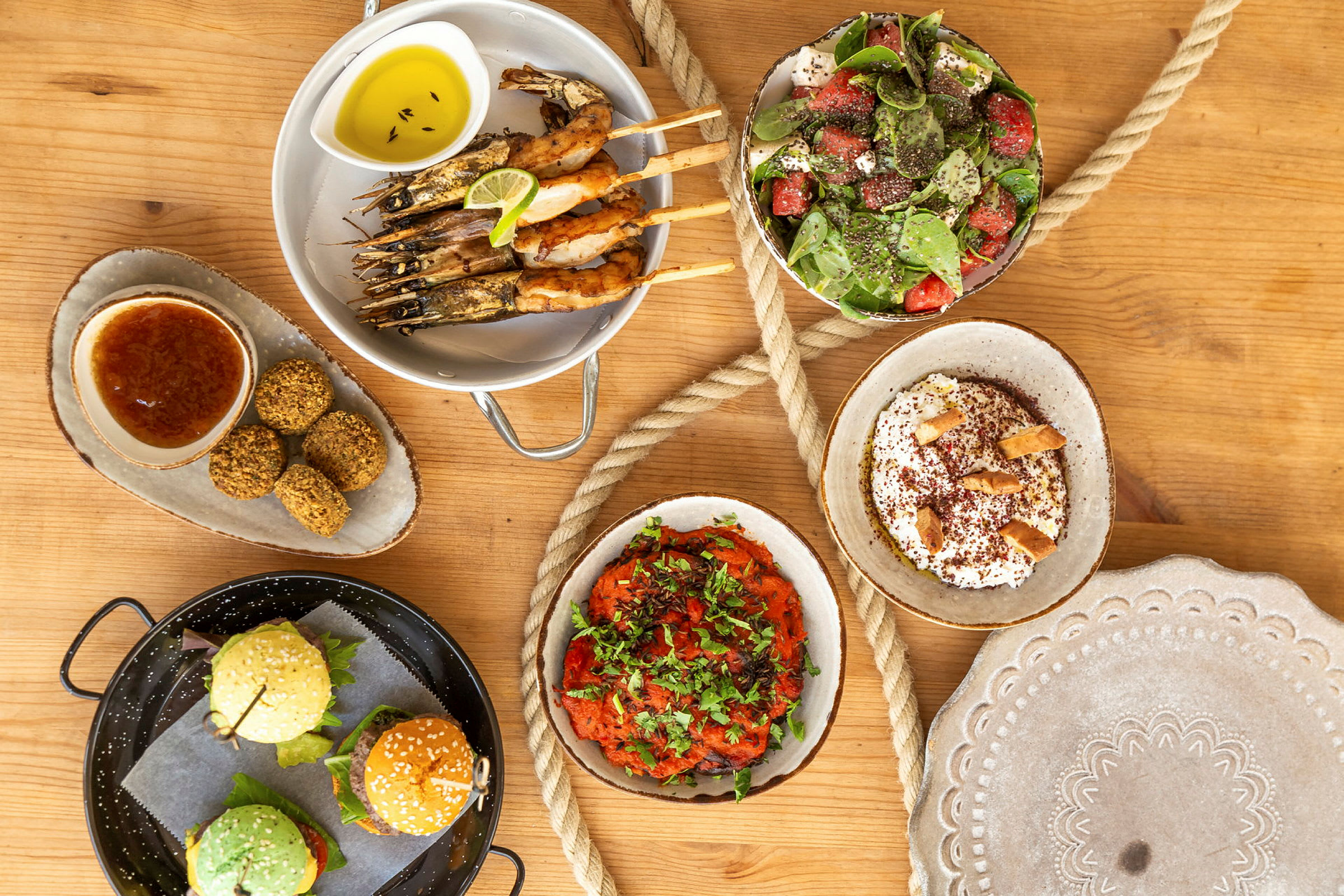 En række forskellige retter såsom rejer på spyd, salat, falafel, burgere, samt dip-saucer og tilbehør på et træbord. (AI-genereret tekst)