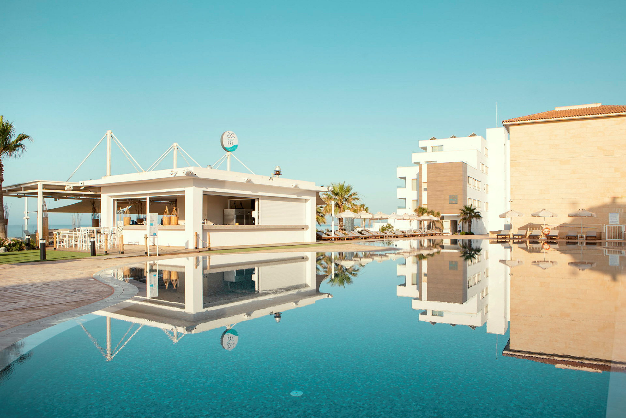 En pool med klart blåt vand omgivet af liggestole og bygninger under en blå himmel. (AI-genereret tekst)