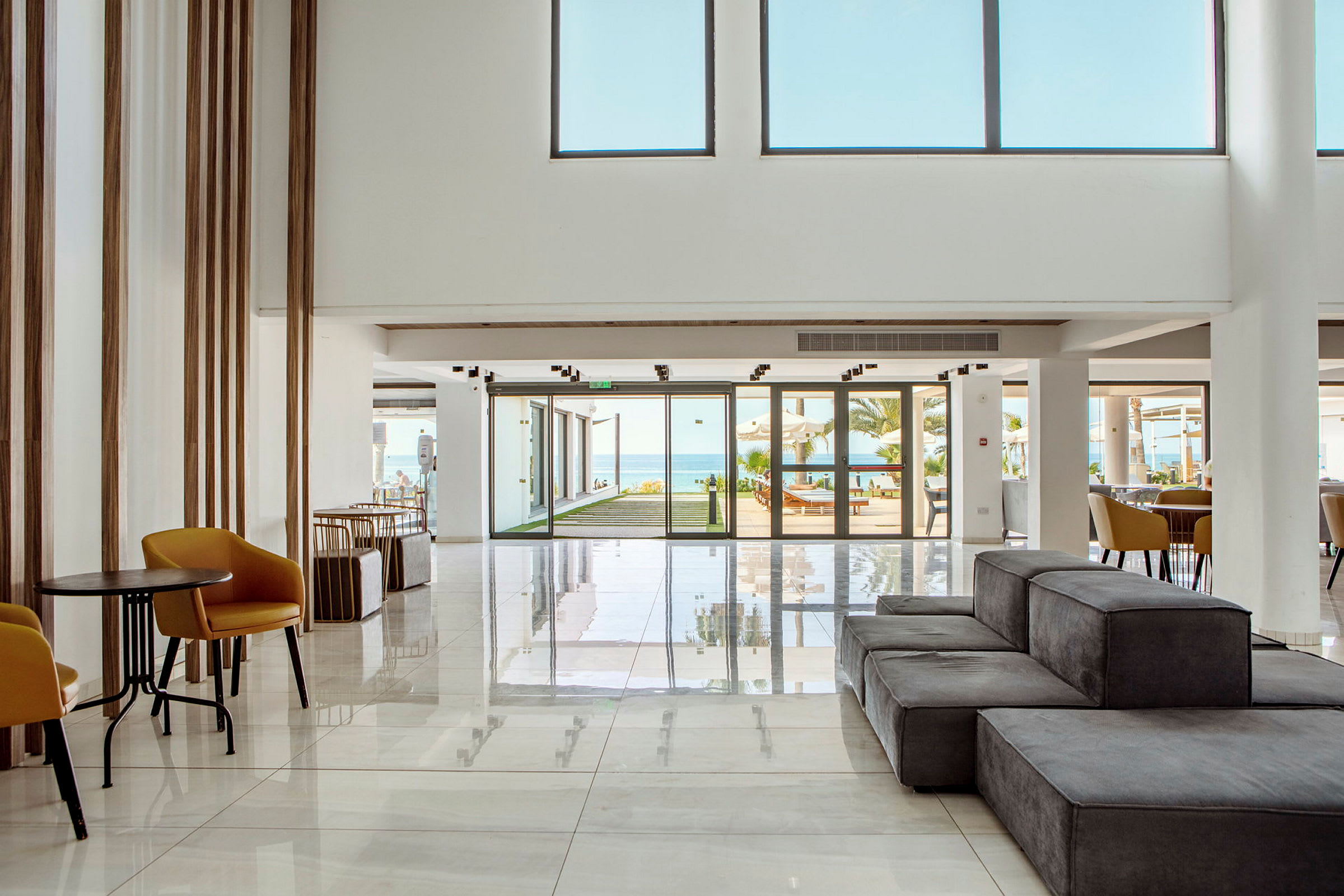 En moderne hotellobby med lys indretning, siddeområder og glasdøre, der fører ud til en udendørs pool og havet. (AI-genereret tekst)