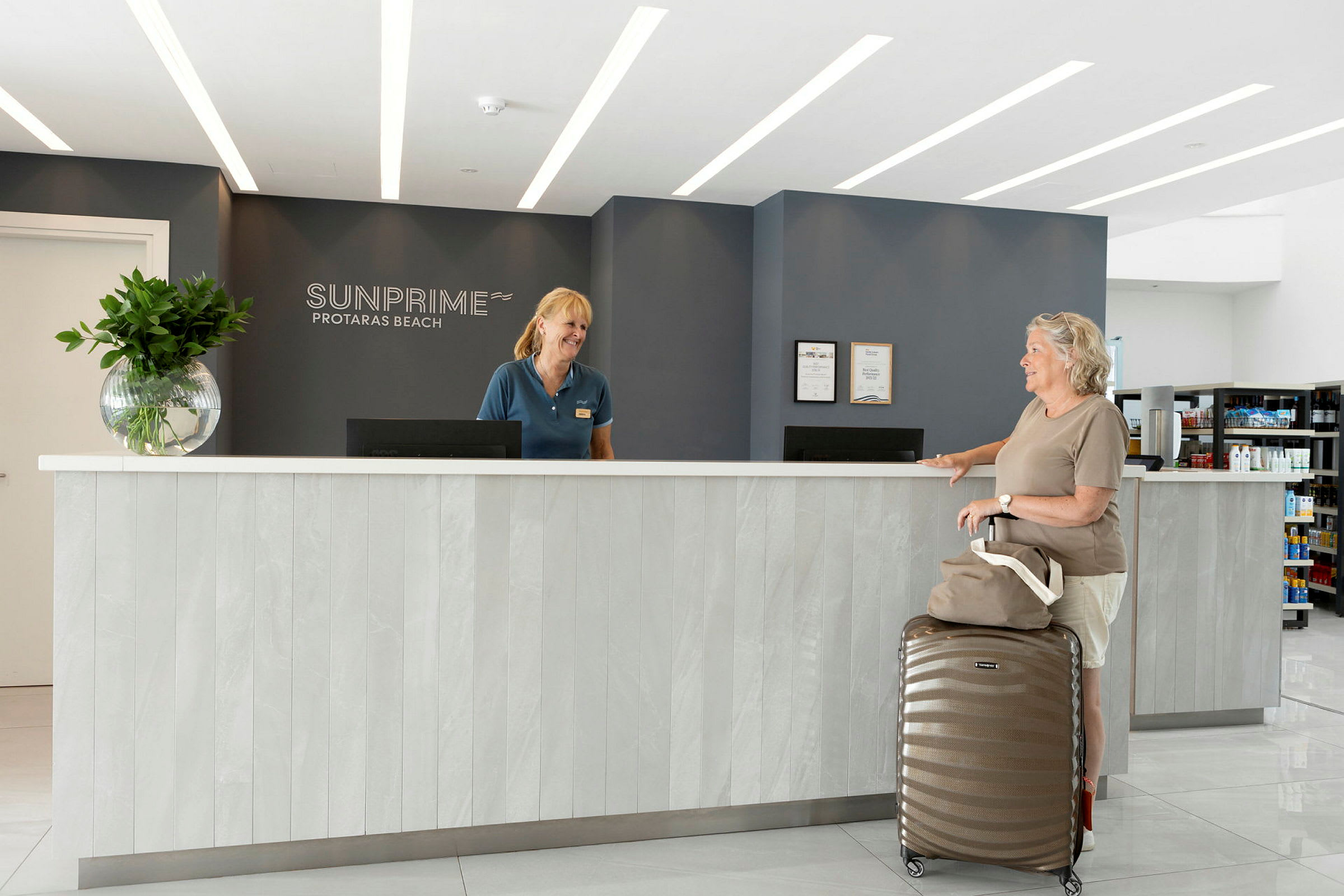 Receptionen på Sunprime Protaras Beach-hotellet med moderne indretning, herunder en skranke med en kuffert og en håndtaske placeret foran. (AI-genereret tekst)