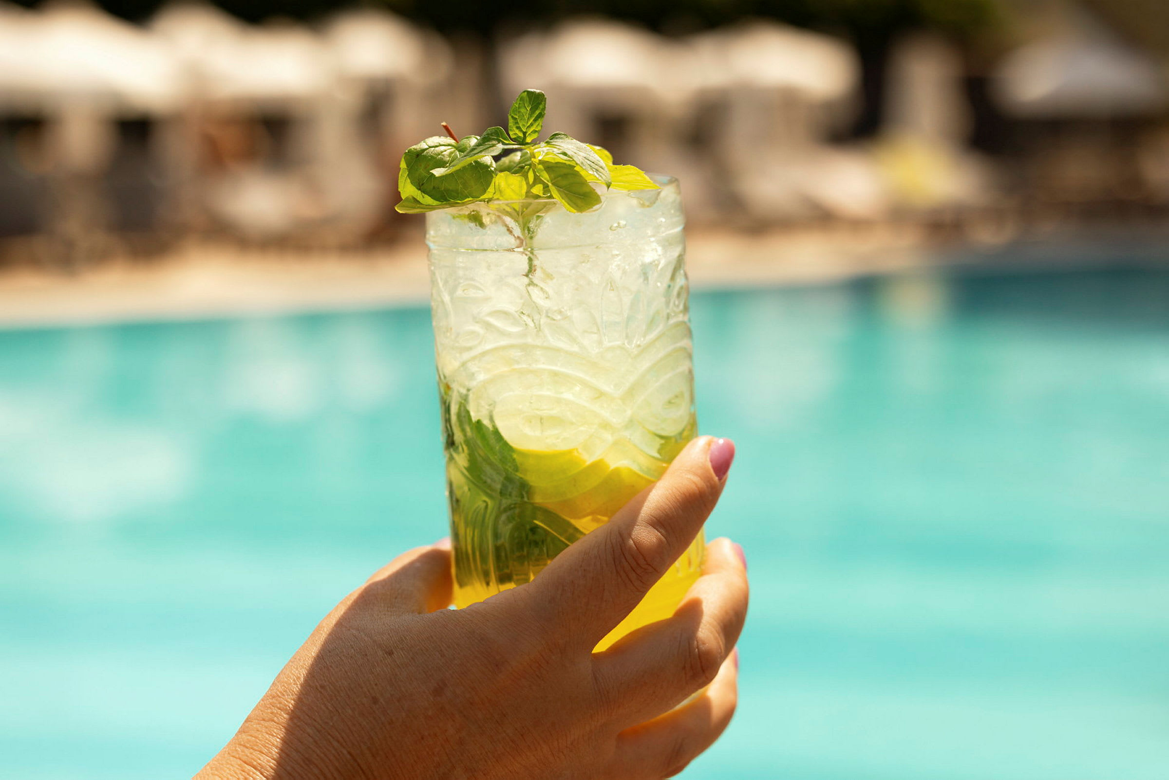 En hånd holder et glas med en kold drik, pyntet med mynte og citron, foran en udendørs swimmingpool. (AI-genereret tekst)