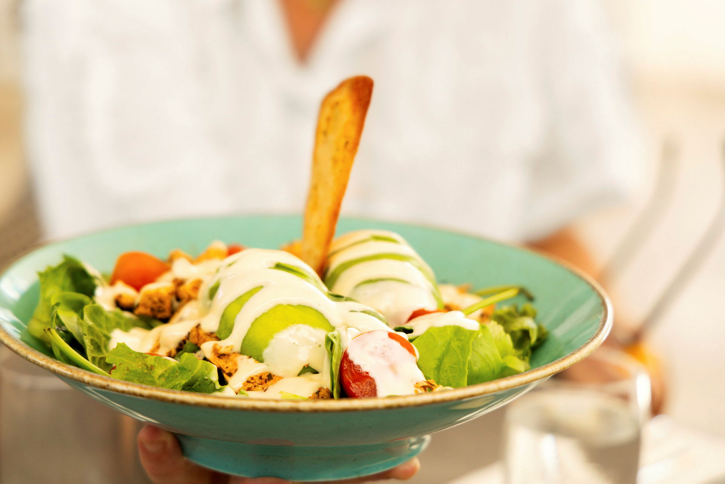 En salat serveret i en turkis skål, indeholdende grøntsager, avocado, dressing og en brødpind. (AI-genereret tekst)
