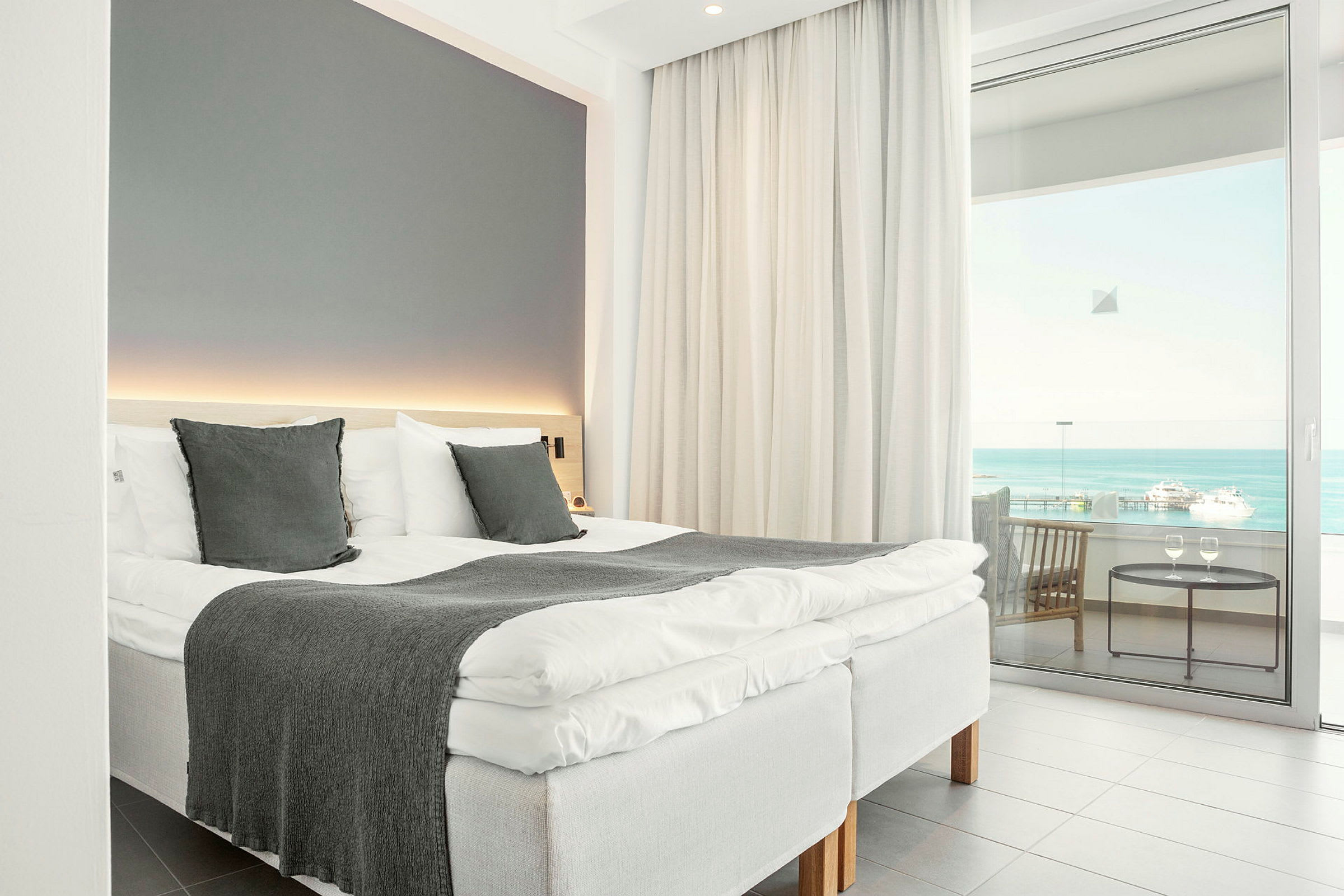 Et hotelværelse med en opredt seng, store vinduer og udsigt over havet. (AI-genereret tekst)