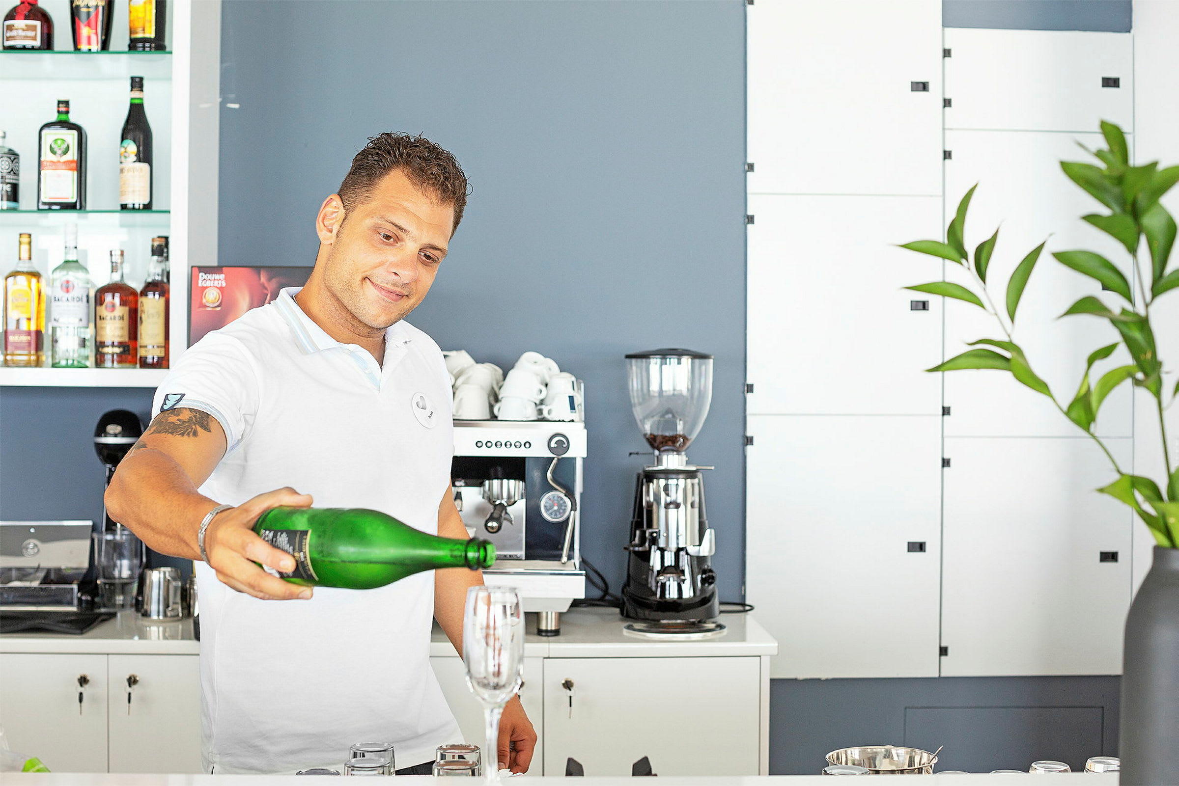 En person holder en grøn flaske og hælder væske i et glas på en bardisk, omgivet af en kaffemaskine og flasker. (AI-genereret tekst)