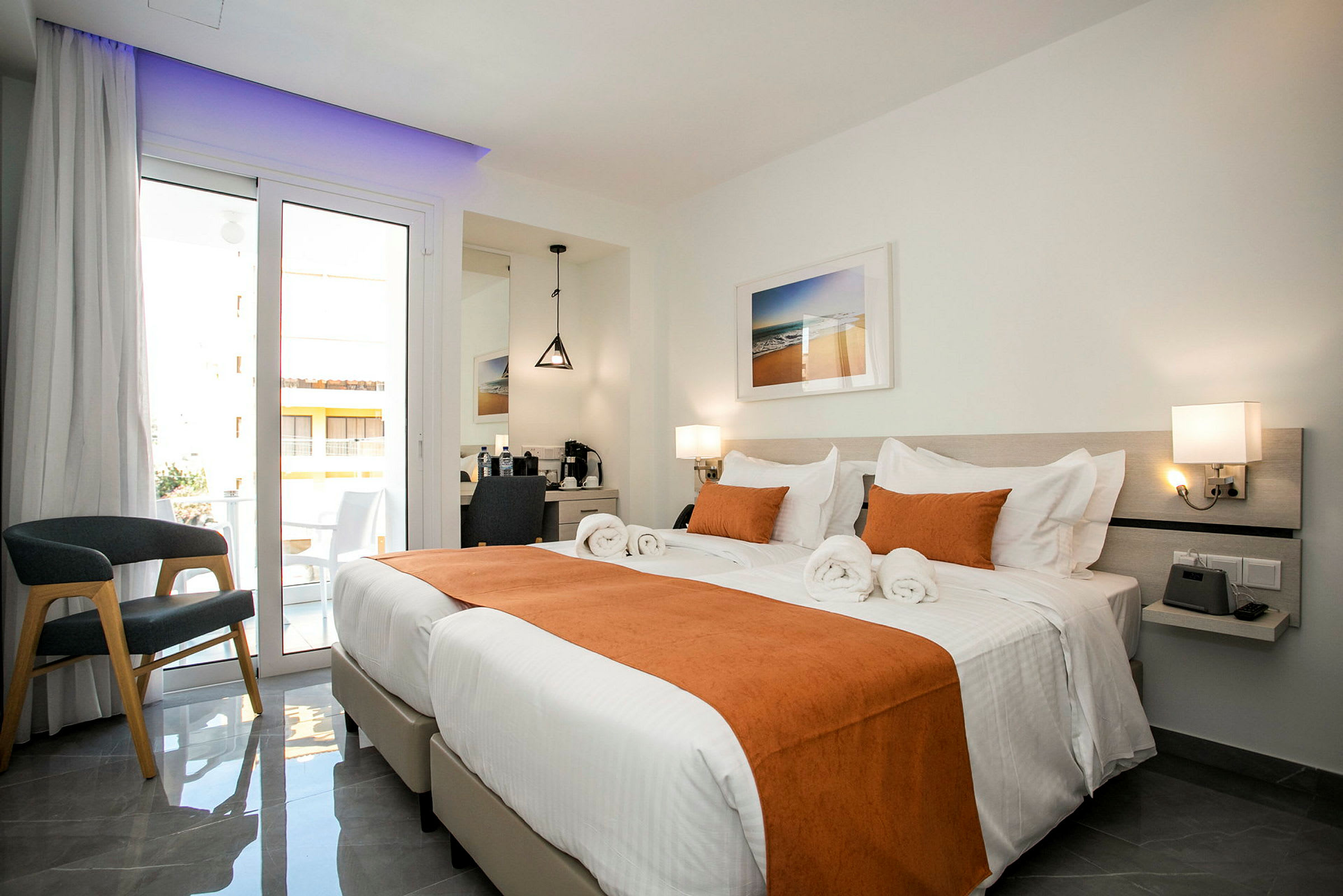 Et lyst hotelværelse med to enkeltsenge redt med hvidt sengetøj og orange udsmykning. Der er en balkon med glasdøre, en stol og et skrivebord med spejl. (AI-genereret tekst)