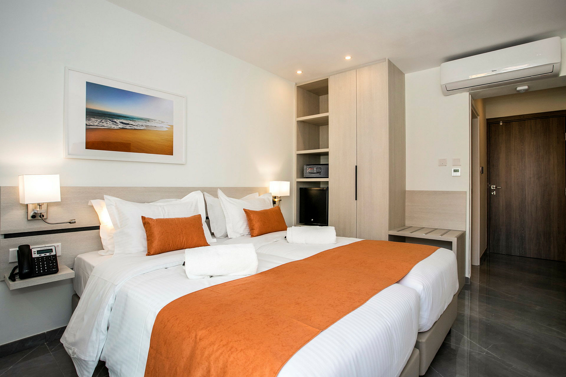Et hotelværelse med en dobbeltseng dækket med hvidt og orange sengetøj. Værelset har moderne møbler, aircondition og vægkunst over sengen. (AI-genereret tekst)