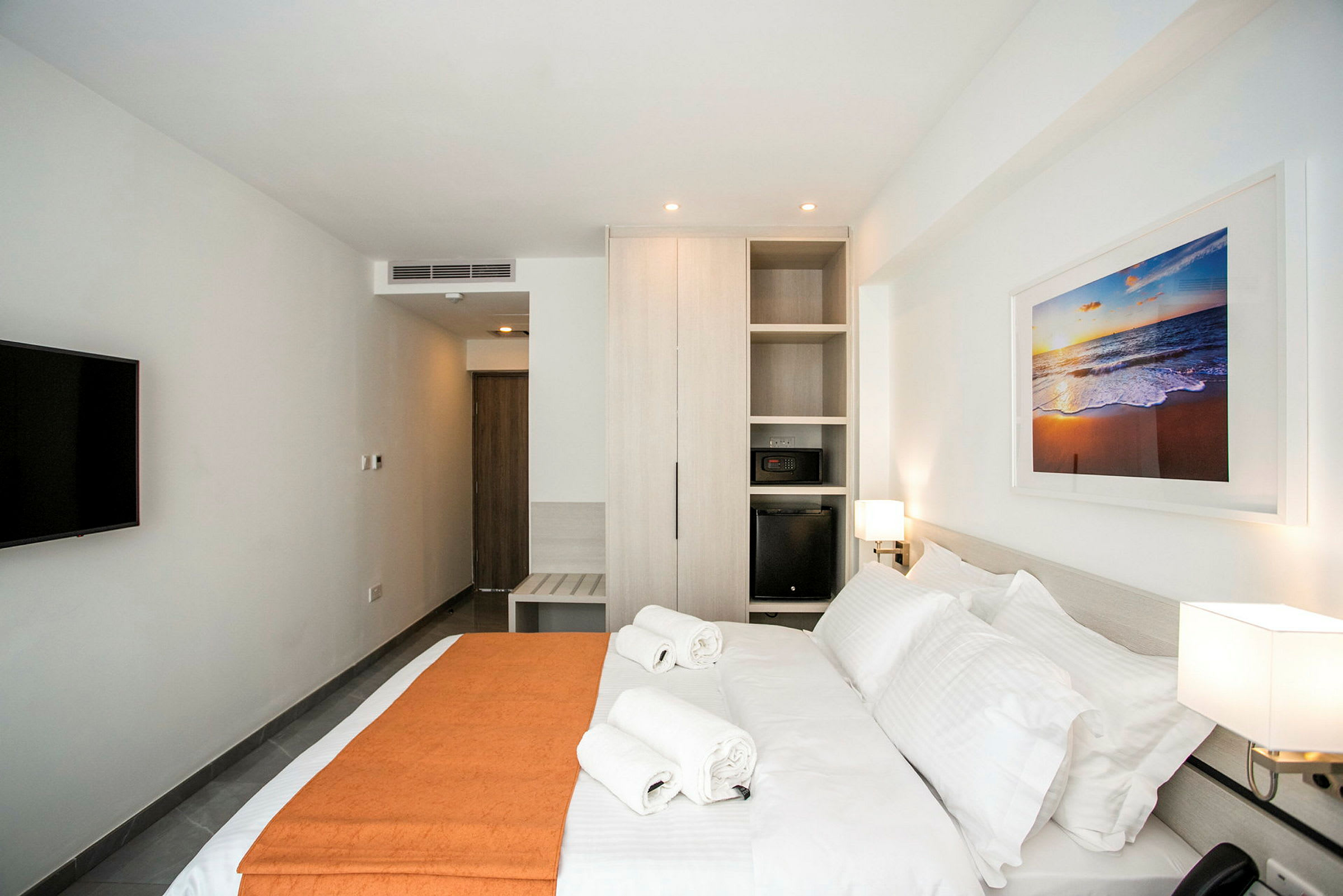 Et moderne hotelværelse med en stor seng, to håndklæder ovenpå, et skab, et tv på væggen og et maleri med motiv af en strand. (AI-genereret tekst)