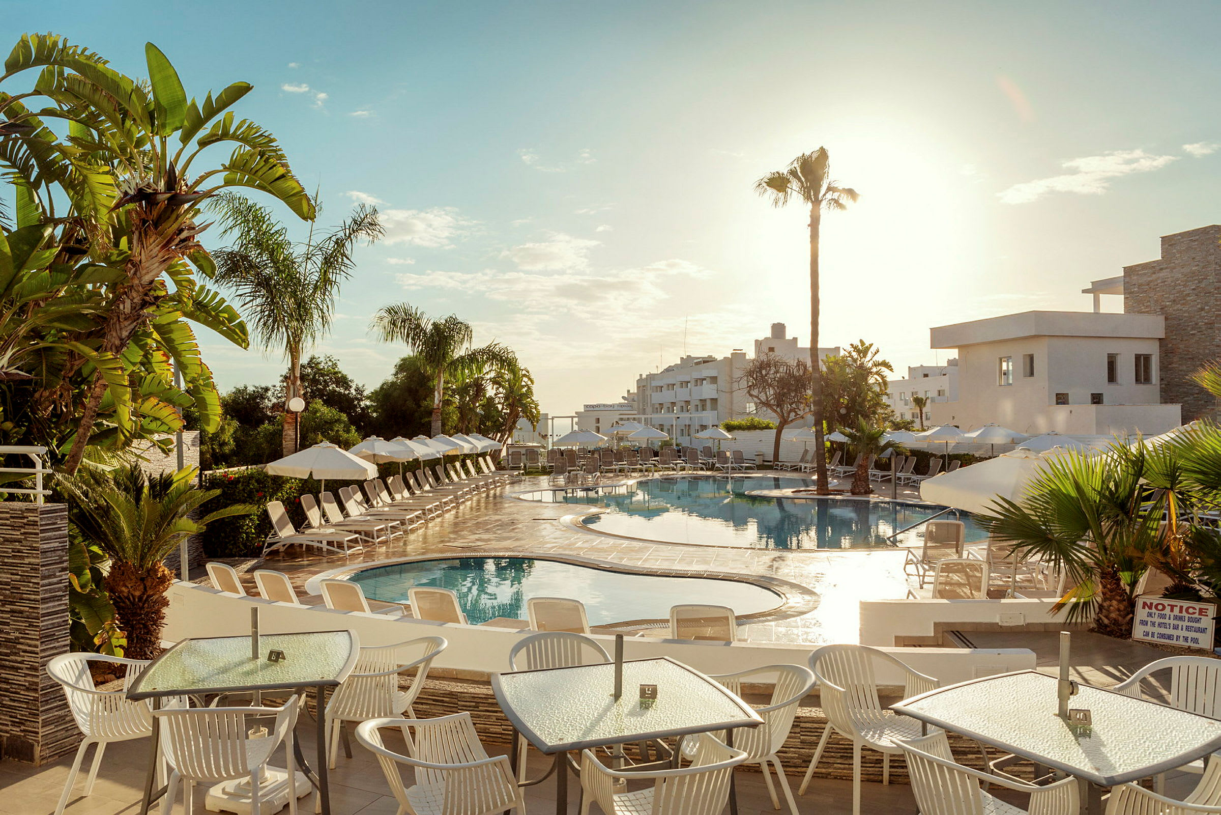 Billedet viser en udendørs swimmingpool på et hotel, omgivet af solsenge og parasoller. I forgrunden er der borde og stole under en solrig himmel. (AI-genereret tekst)
