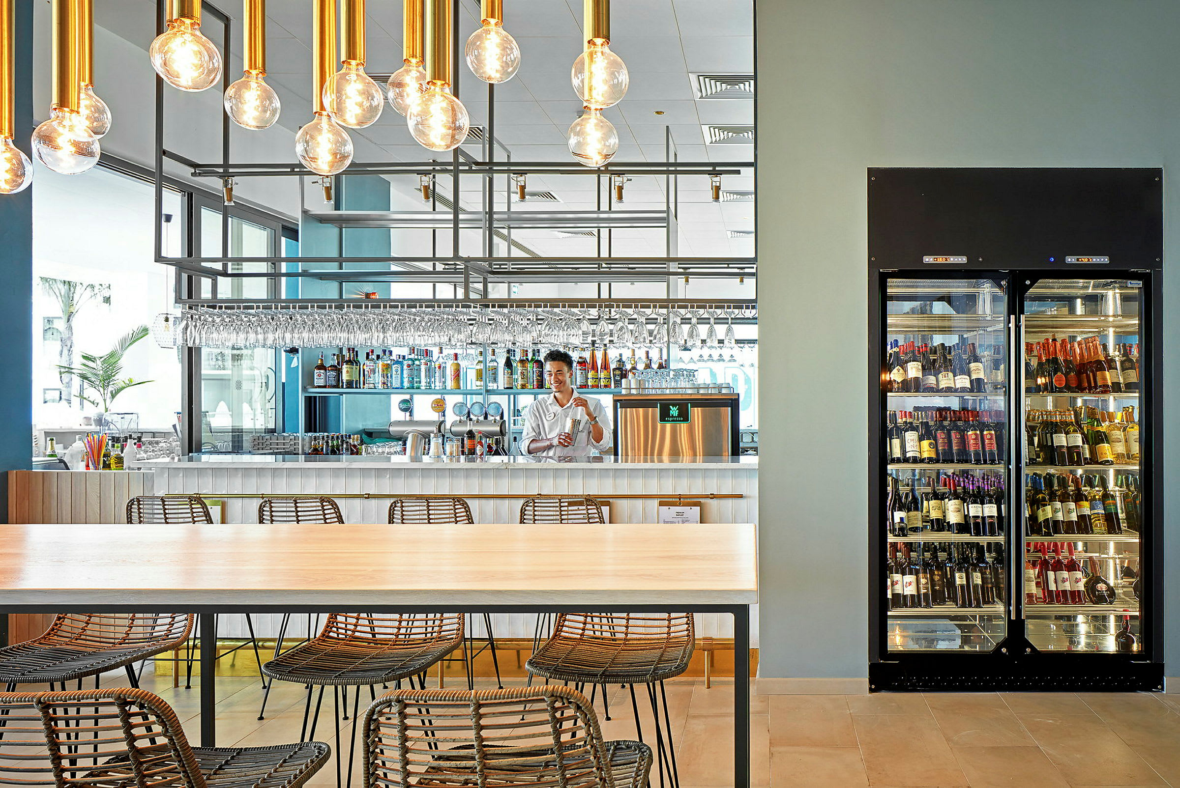 Restaurantinteriør med en bar, rummeligt bord og stole samt et køleskab med flasker. (AI-genereret tekst)