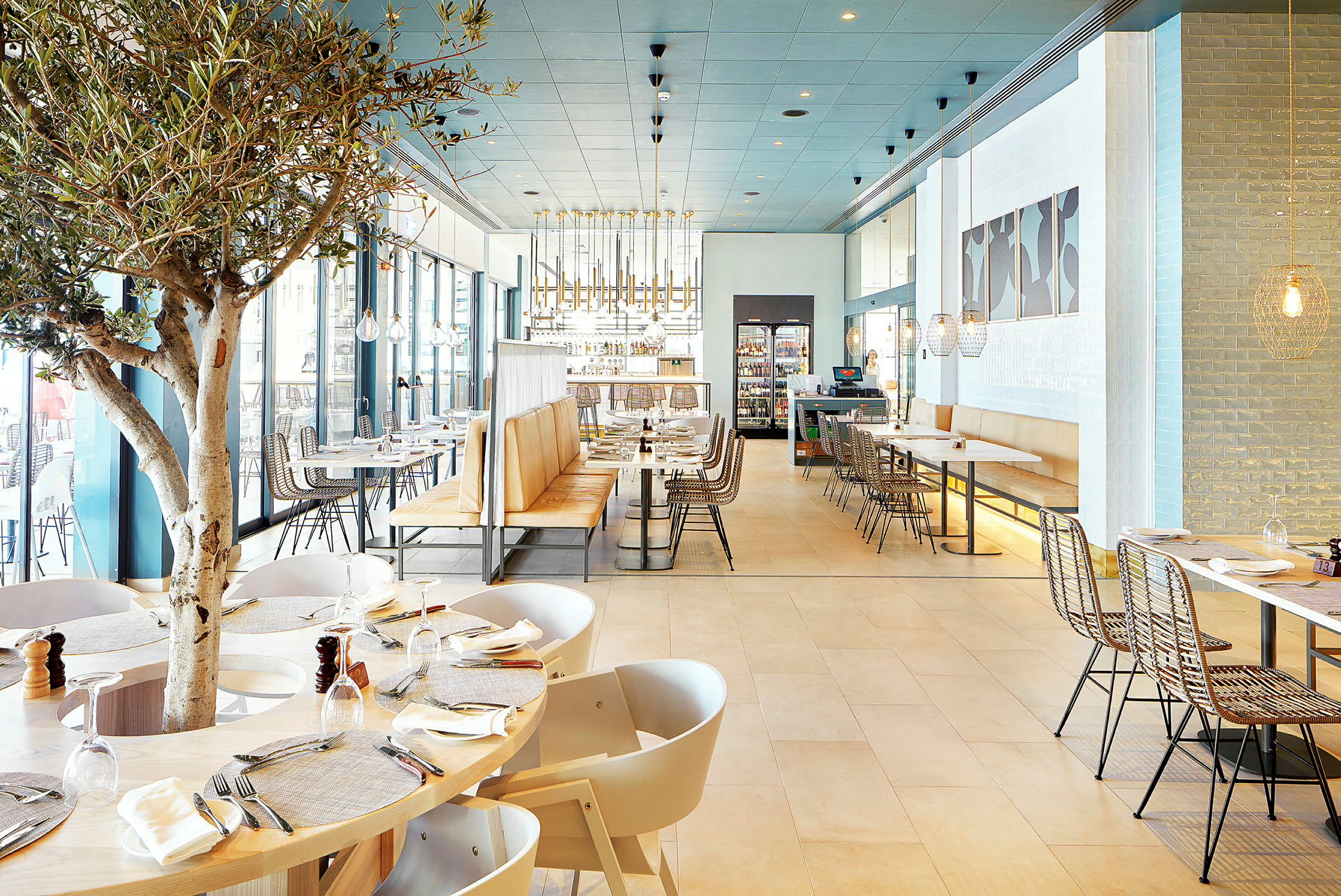 Interiør af en moderne restaurant med træmøbler, lyse farver og et træ midt blandt bordene. (AI-genereret tekst)