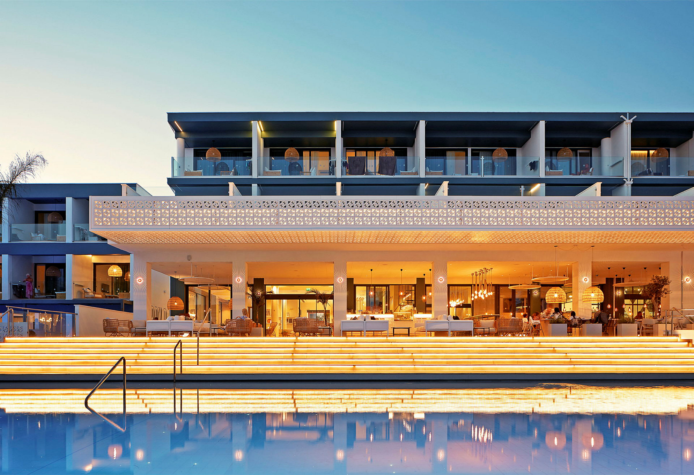 Hotellets facade med oplyste trapper og en pool i forgrunden om aftenen. (AI-genereret tekst)