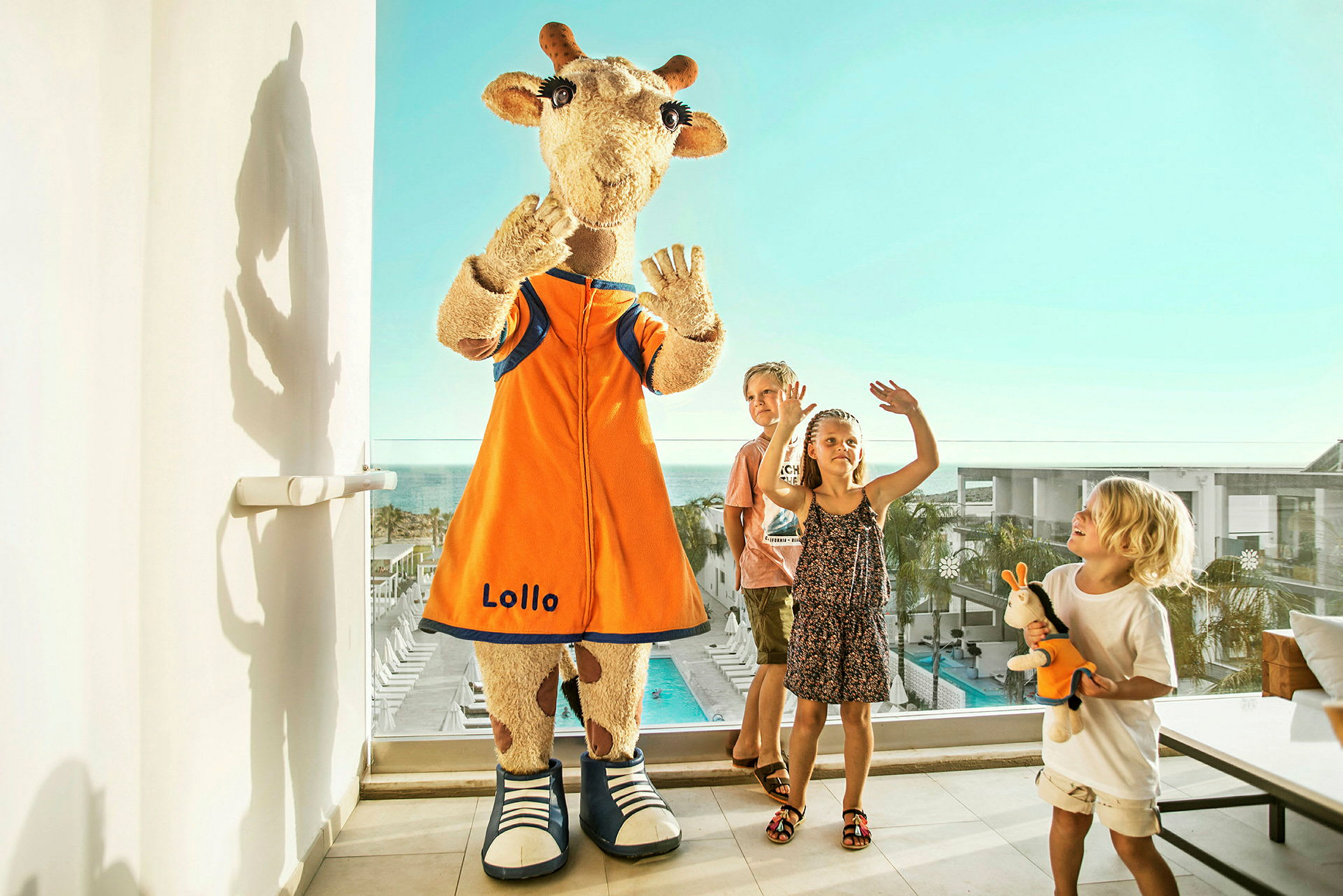 Maskotten Lollo står på en terrasse nær glaspartier med udsigt over en pool og omkringliggende bygninger. (AI-genereret tekst)
