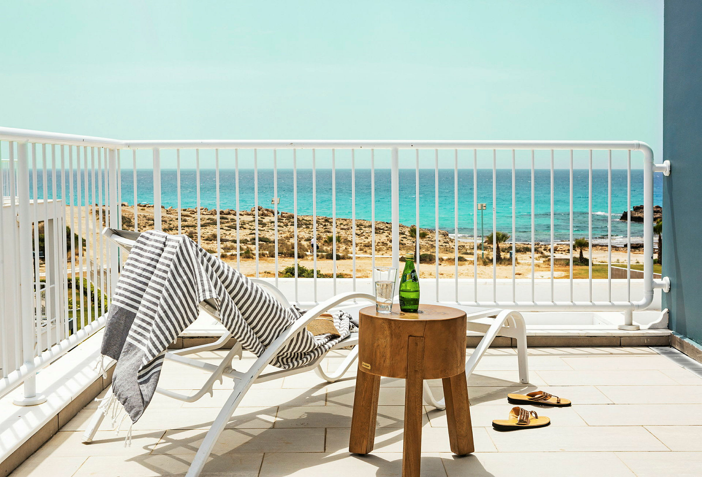 En liggestol står på en terrasse med udsigt over havet, et stribet håndklæde hænger på stolen, og et lille træbord med en flaske og et glas står ved siden af. (AI-genereret tekst)
