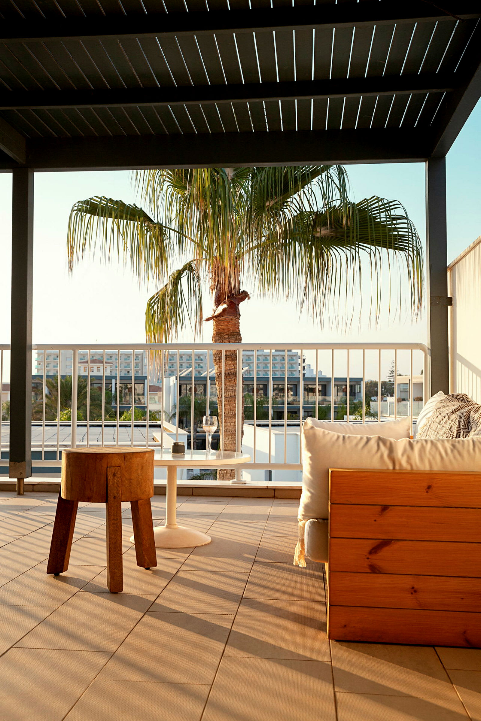 Terrasse med træstol, hvidt bord og træbænk, en palme ses bag rækværket. (AI-genereret tekst)