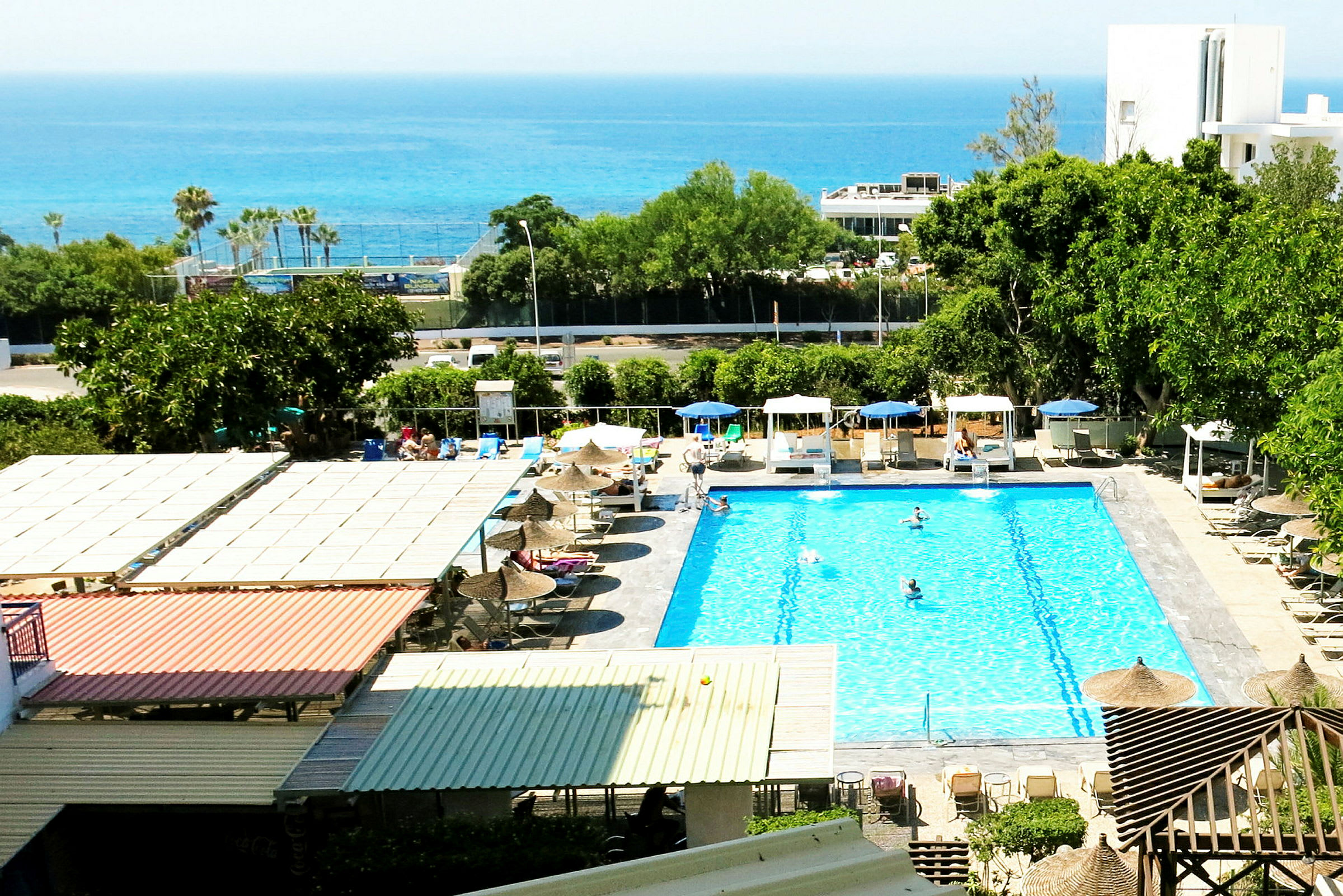 Billede af en udendørs swimmingpool omgivet af liggestole og parasoller, med havet i baggrunden. (AI-genereret tekst)