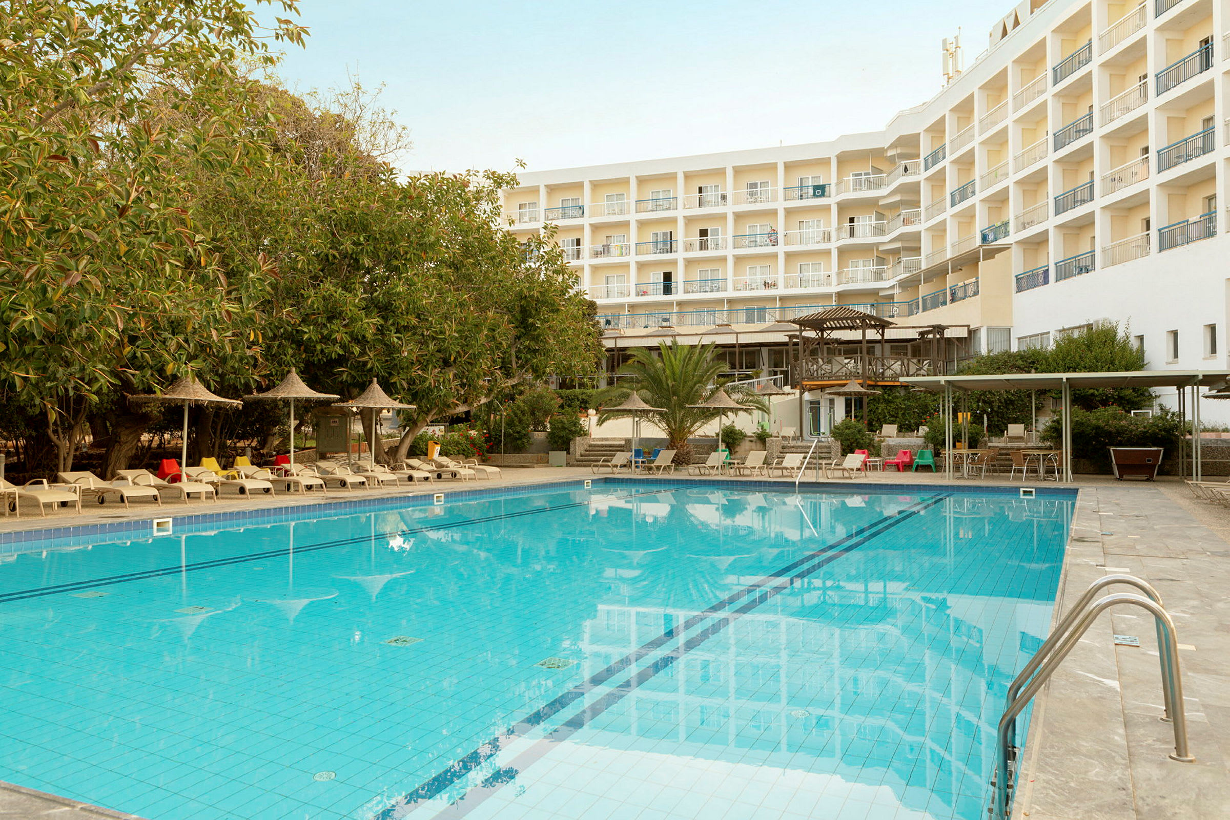 En udendørs pool omgivet af liggestole, parasoller og et hotel med flere etager i baggrunden. (AI-genereret tekst)