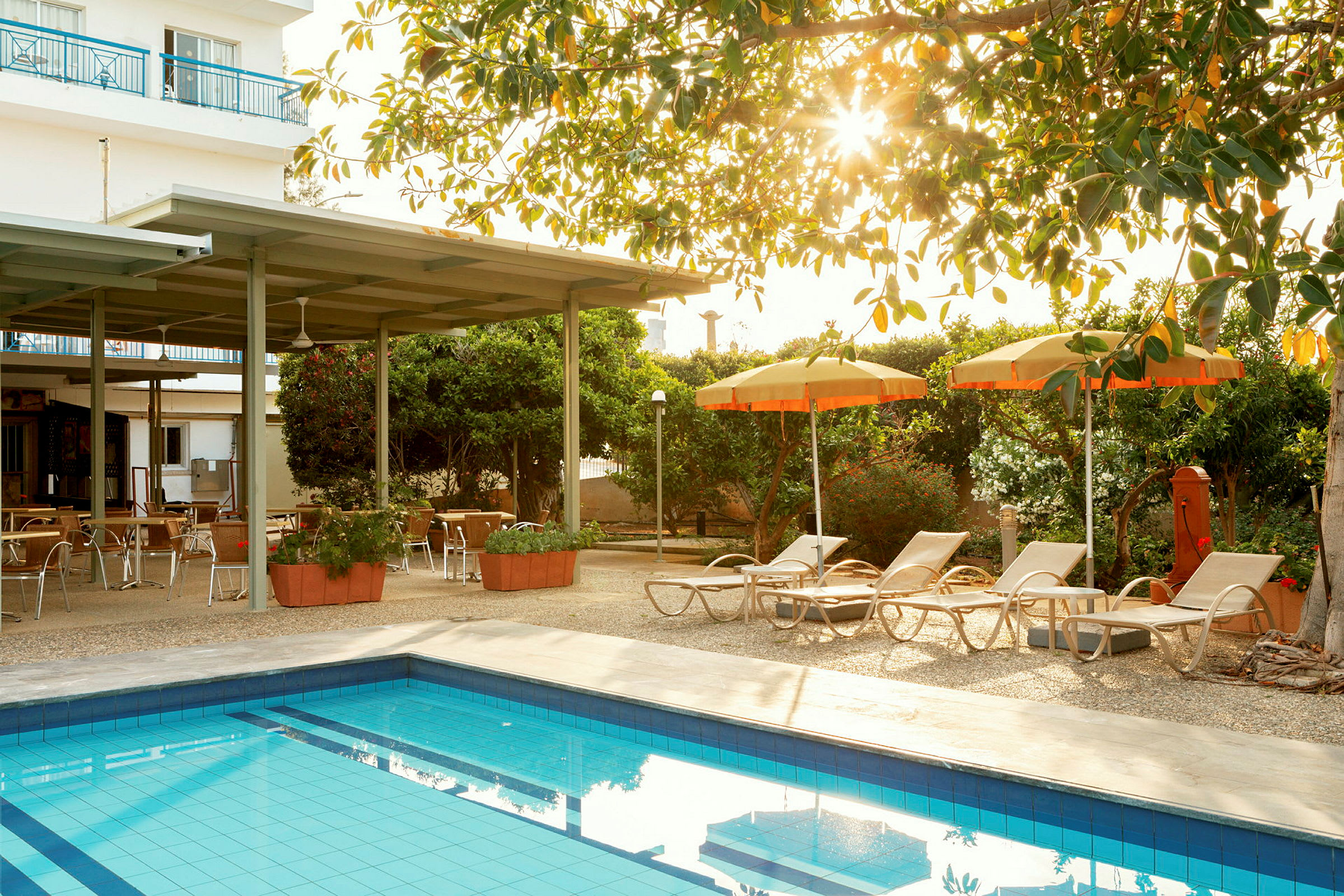 Udendørsområde med en pool, liggestole og parasoller samt siddepladser under en pergola. (AI-genereret tekst)