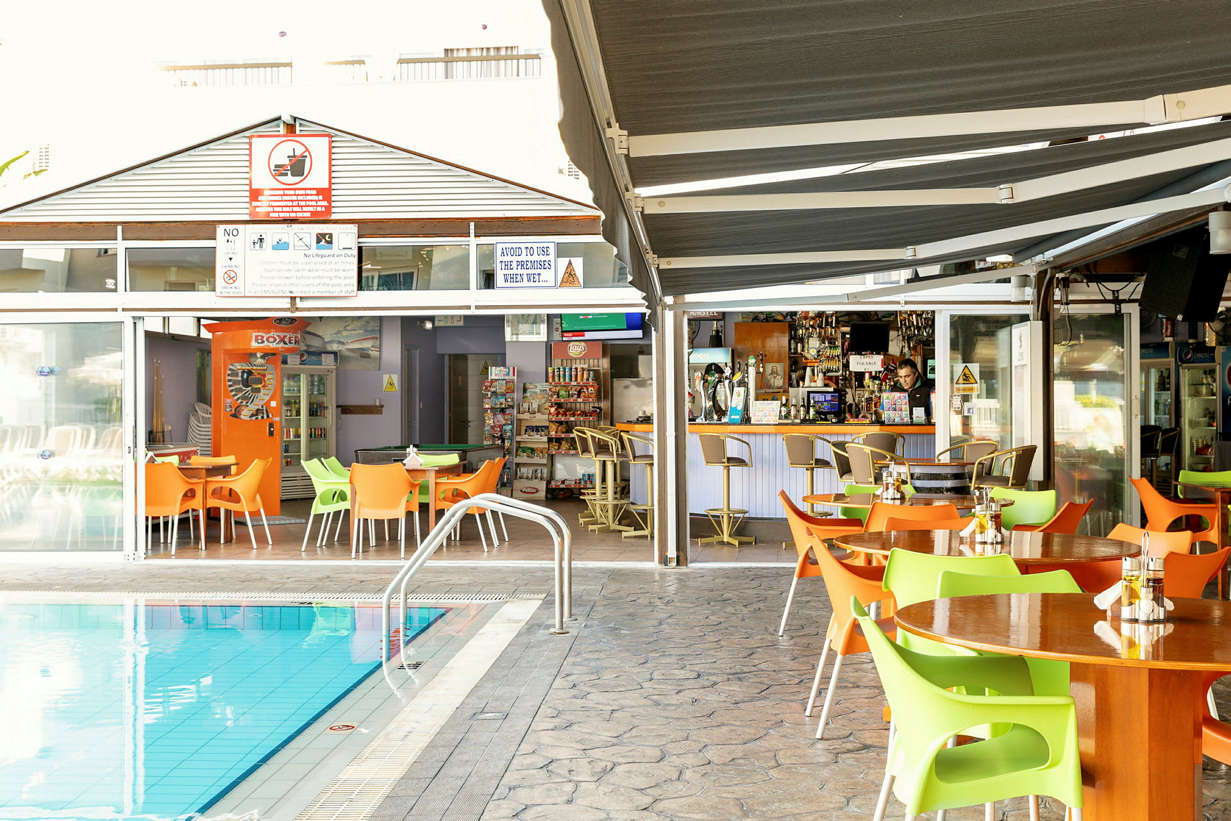 Udendørsområde med borde og stole ved en pool, med en bar og spisesteder i baggrunden. (AI-genereret tekst)