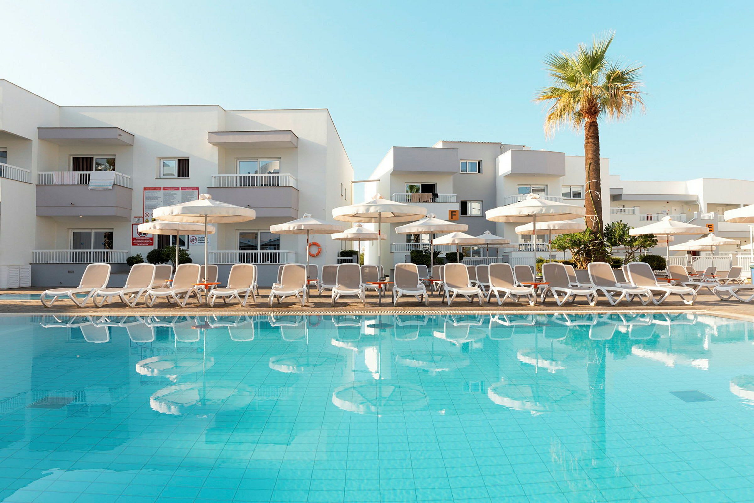 Billede af en pool med liggestole og parasoller foran et hotel. (AI-genereret tekst)