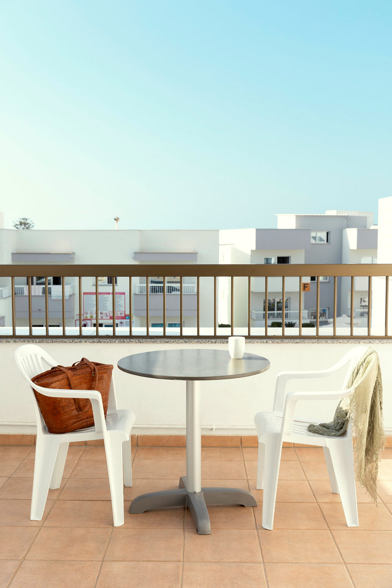 To hvide plastikstole og et bord på en terrasse med rækværk og moderne bygningsarkitektur i baggrunden. (AI-genereret tekst)