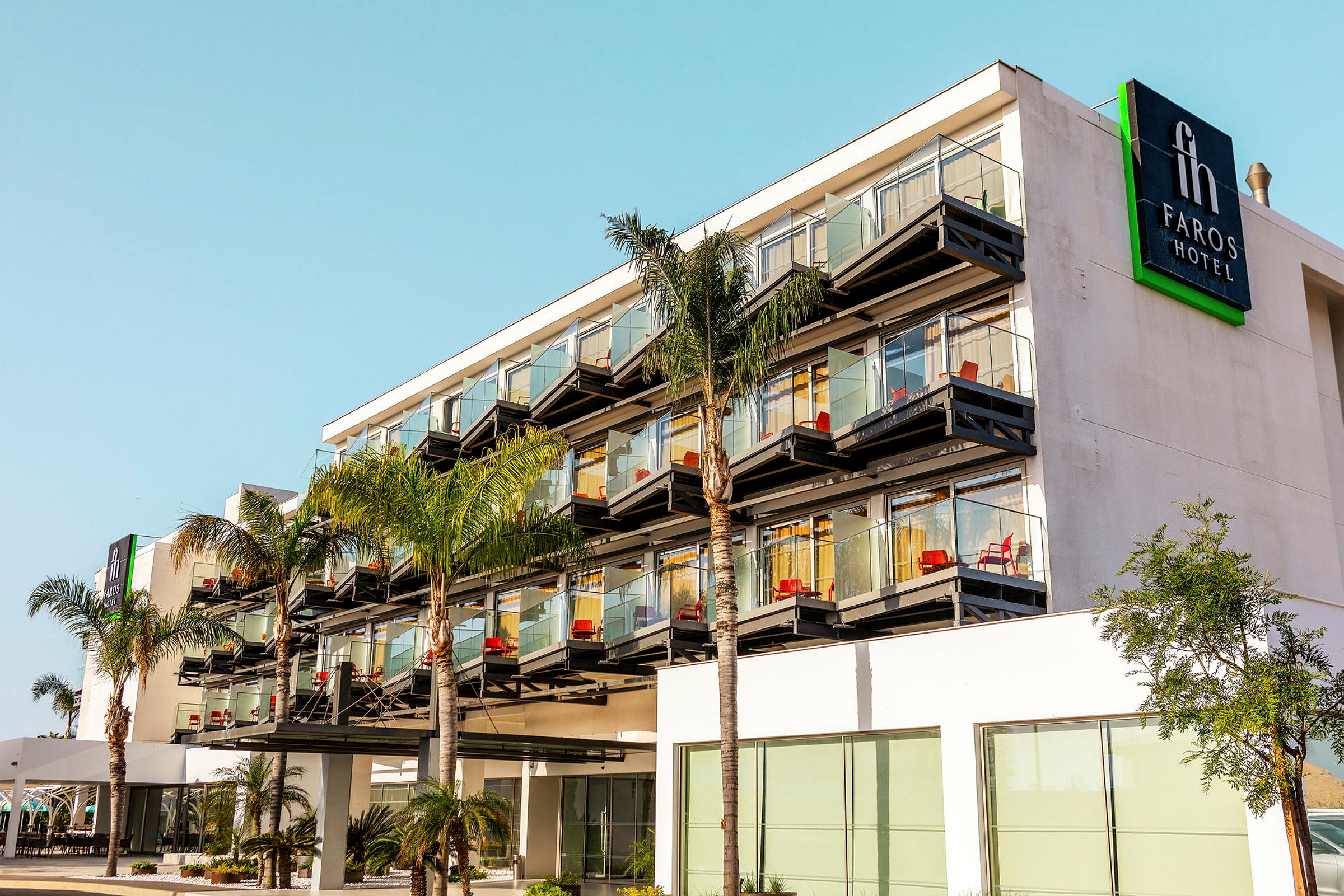 Udendørs billede af Faros Hotel med flere etager, glasaltaner og palmetræer foran. (AI-genereret tekst)