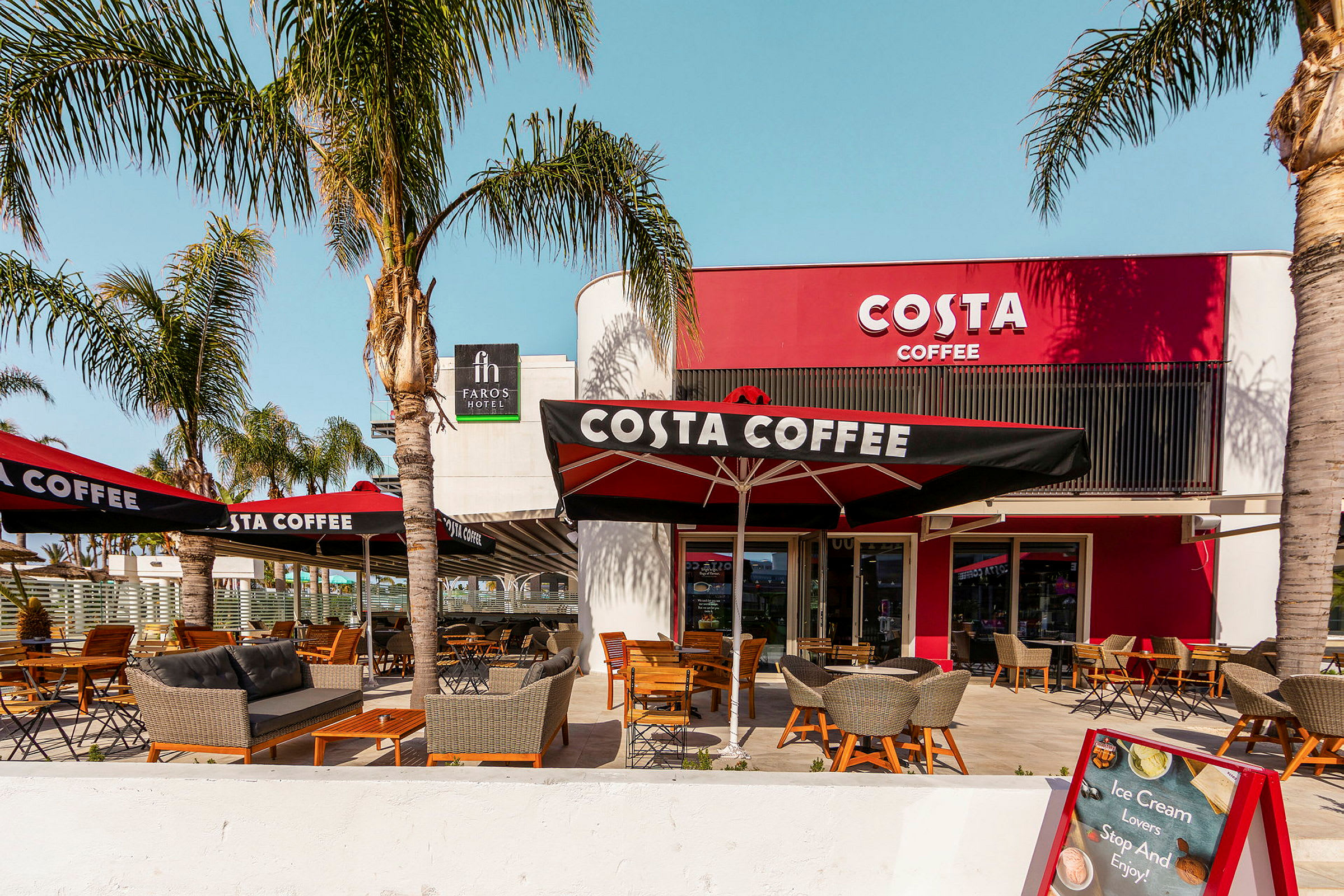 Udendørs serveringsområde ved caféen Costa Coffee med palmer og siddepladser. (AI-genereret tekst)