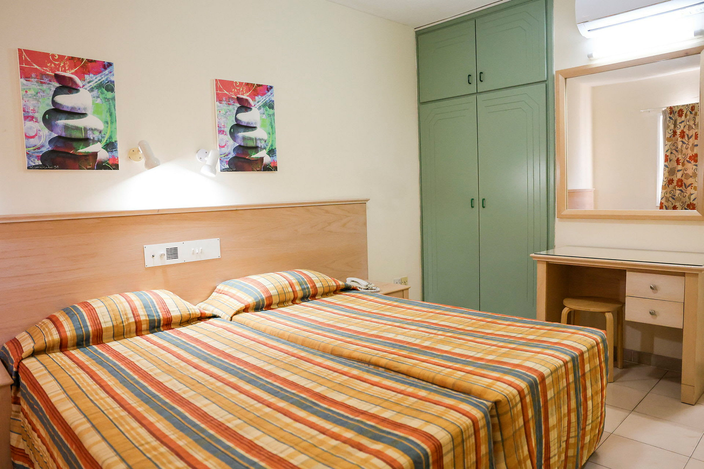 Hotelværelse med to enkeltsenge og ternede sengetæpper. Der er et grønt skab, et spejl over et skrivebord og kunstmalerier på væggen. (AI-genereret tekst)