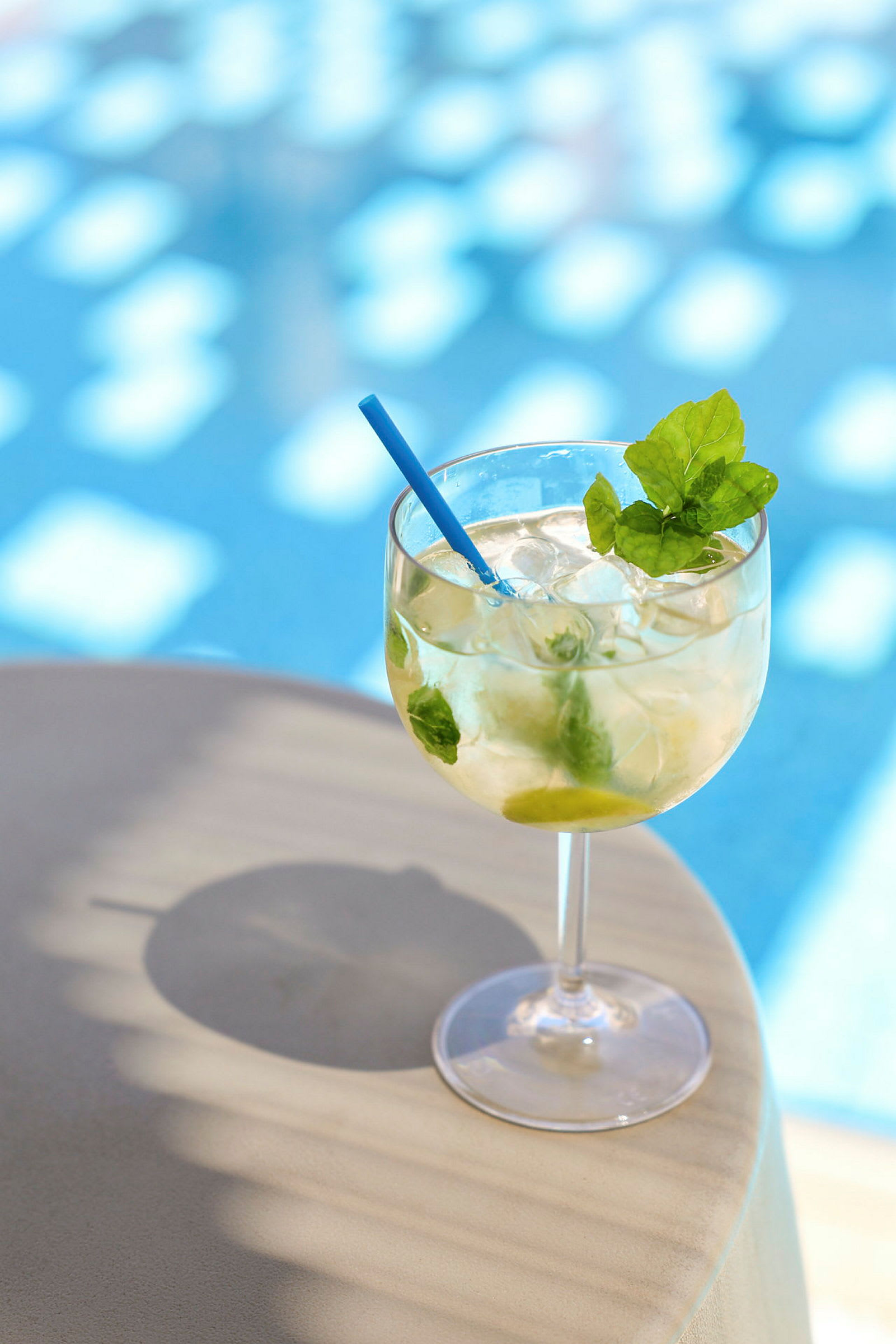 Et glas med en kold drik, dekoreret med et blå sugerør og frisk mynte, står på et bord tæt på en swimmingpool. (AI-genereret tekst)