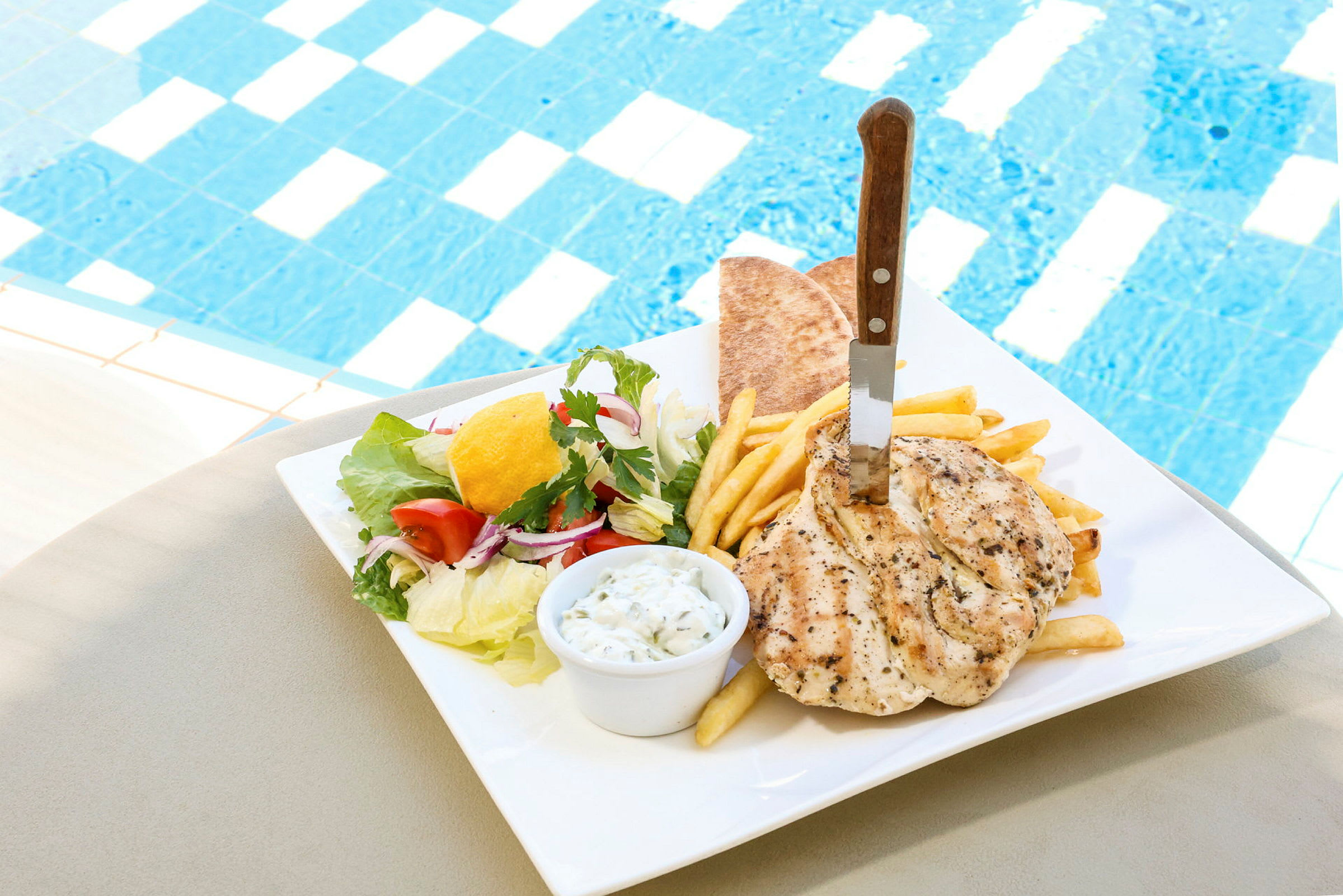 En tallerken med kylling, pommes frites, pitabrød og salat ved siden af en pool. (AI-genereret tekst)