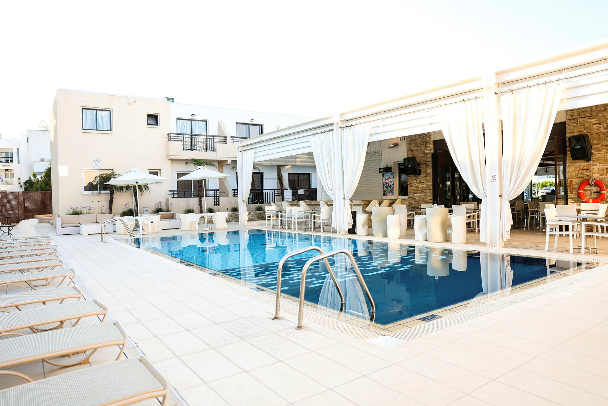 Et hotelområde med en pool omgivet af liggestole og hvide bygninger med altaner. (AI-genereret tekst)