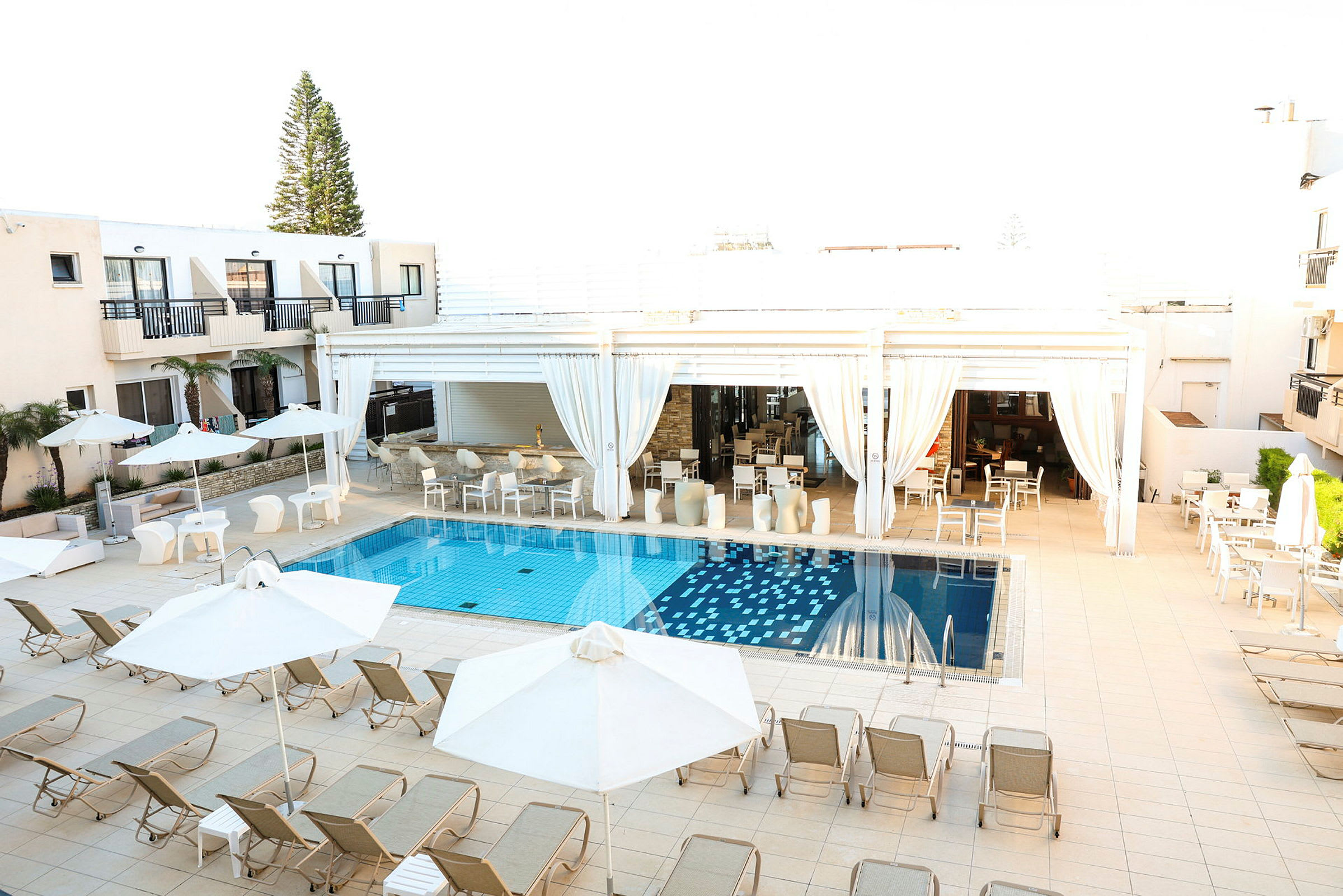 Udendørs swimmingpool med liggestole og parasoller, omgivet af lyse bygninger og siddeområder. (AI-genereret tekst)