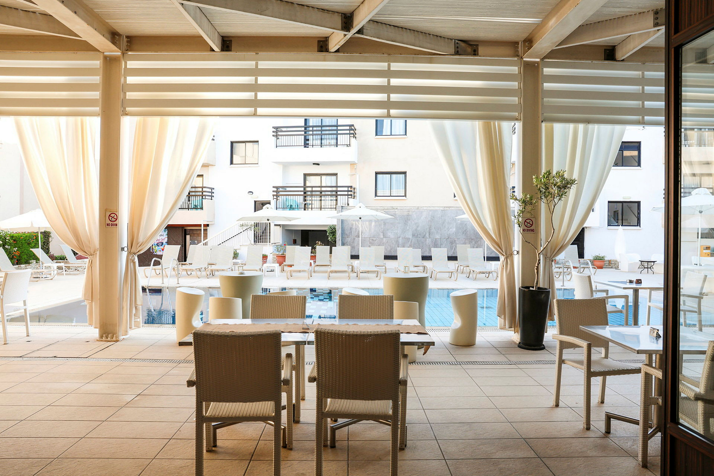 En udendørs terrasse med borde og stole under en overdækket struktur med udsigt til en pool med liggestole og parasoller. (AI-genereret tekst)