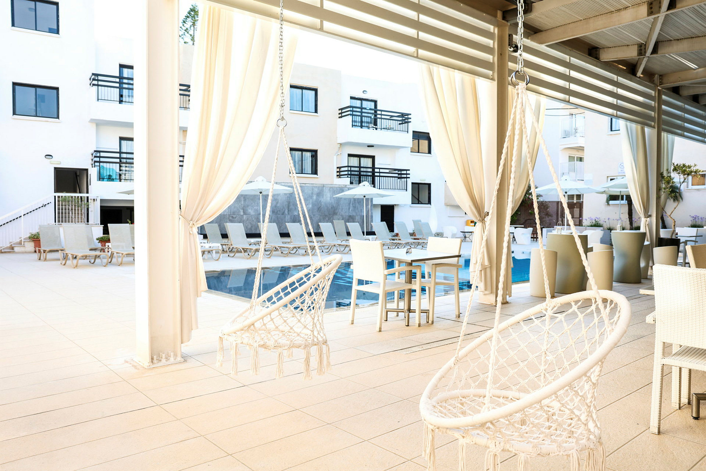 Billede af en solrig hotelpool med liggestole og parasoller samt to hvide hængestole under et tag. (AI-genereret tekst)
