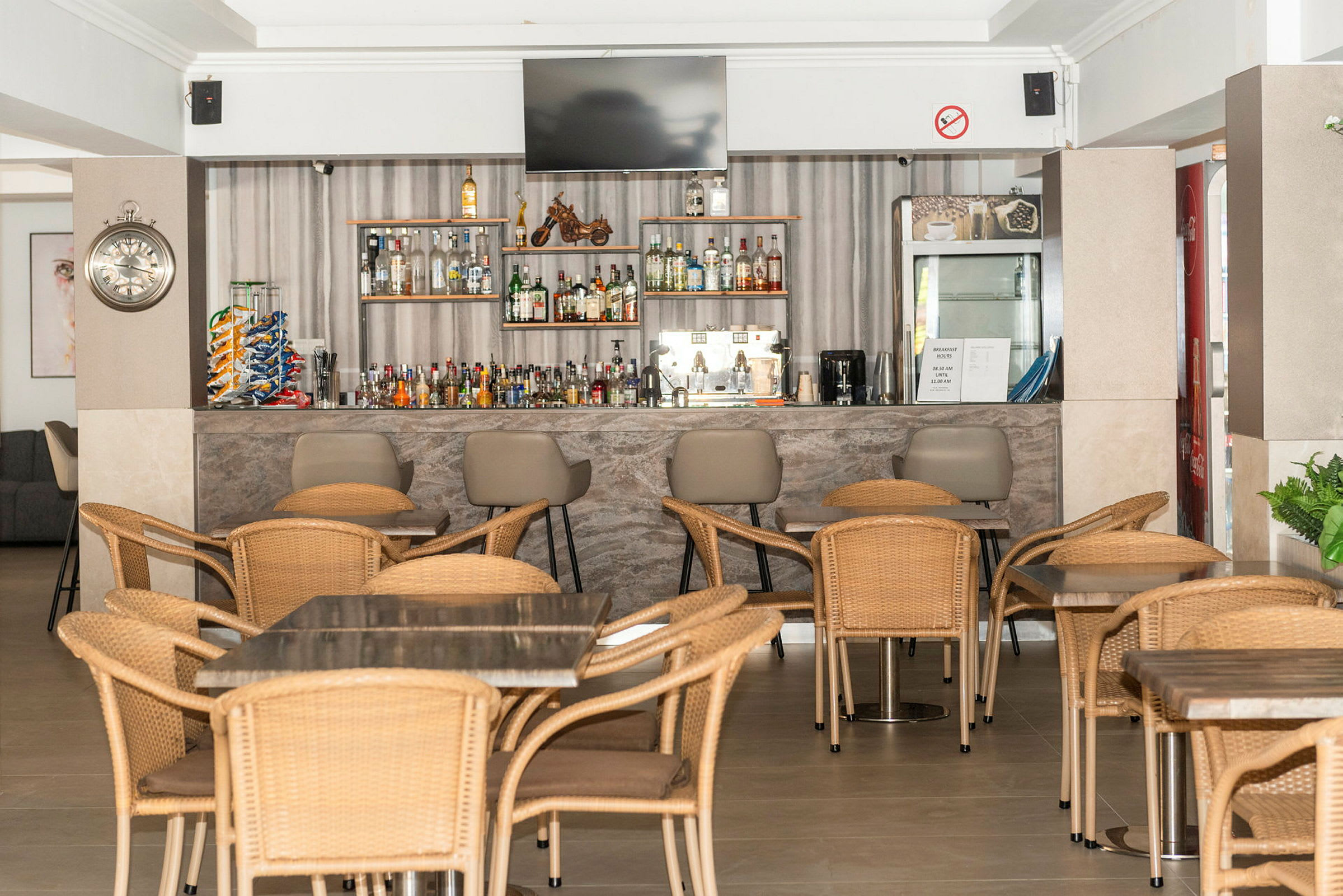 En hotelbar med borde og stole af rattan samt en velassorteret hylde med drikkevarer. (AI-genereret tekst)