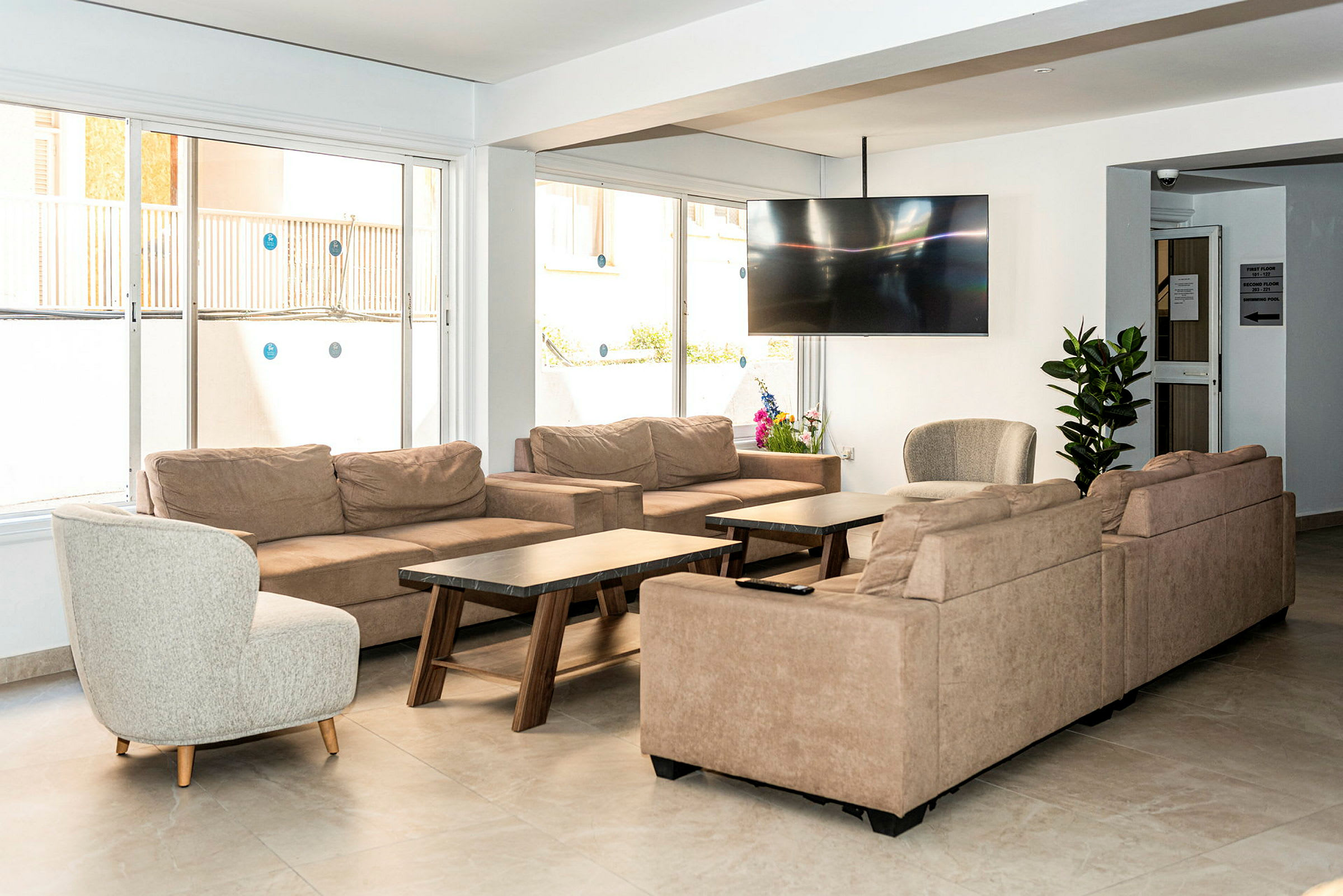 En dagligstue med beige sofaer, en rund lænestol, sofaborde og et stort fladskærms-tv på væggen samt planter som dekoration. (AI-genereret tekst)