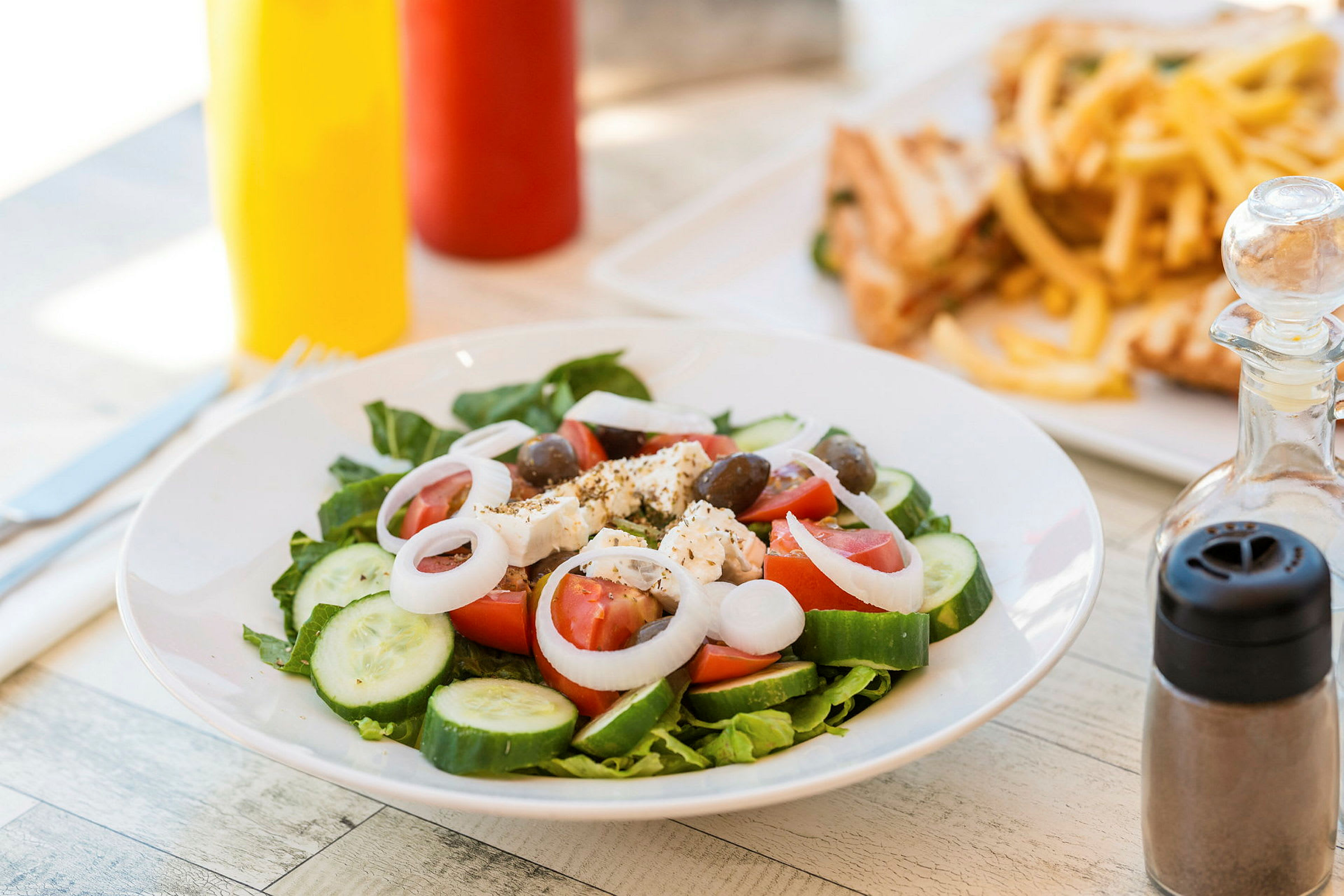 En tallerken med salat indeholdende tomat, agurk, løg, fetaost og oliven. I baggrunden ses pommes frites og sandwich. (AI-genereret tekst)
