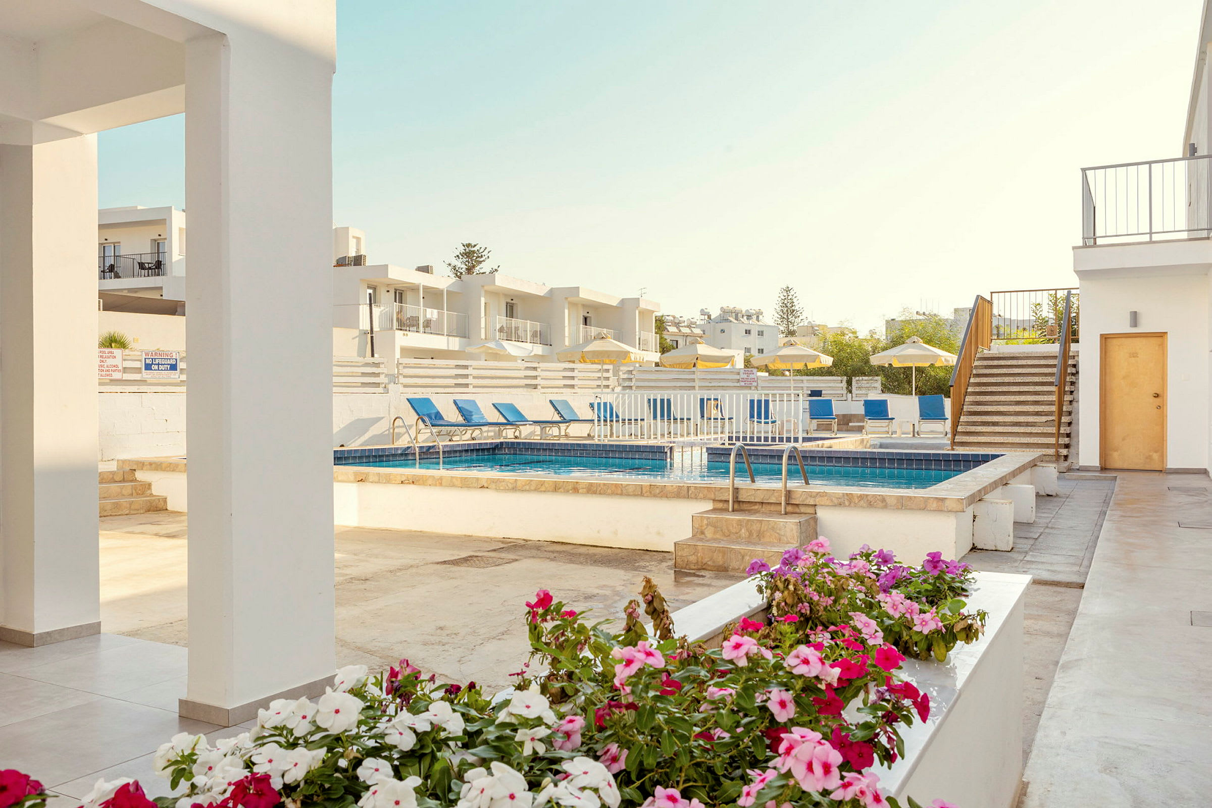 Billede af en hotelpool med liggestole og parasoller, omgivet af hvide bygninger og blomster i forgrunden. (AI-genereret tekst)
