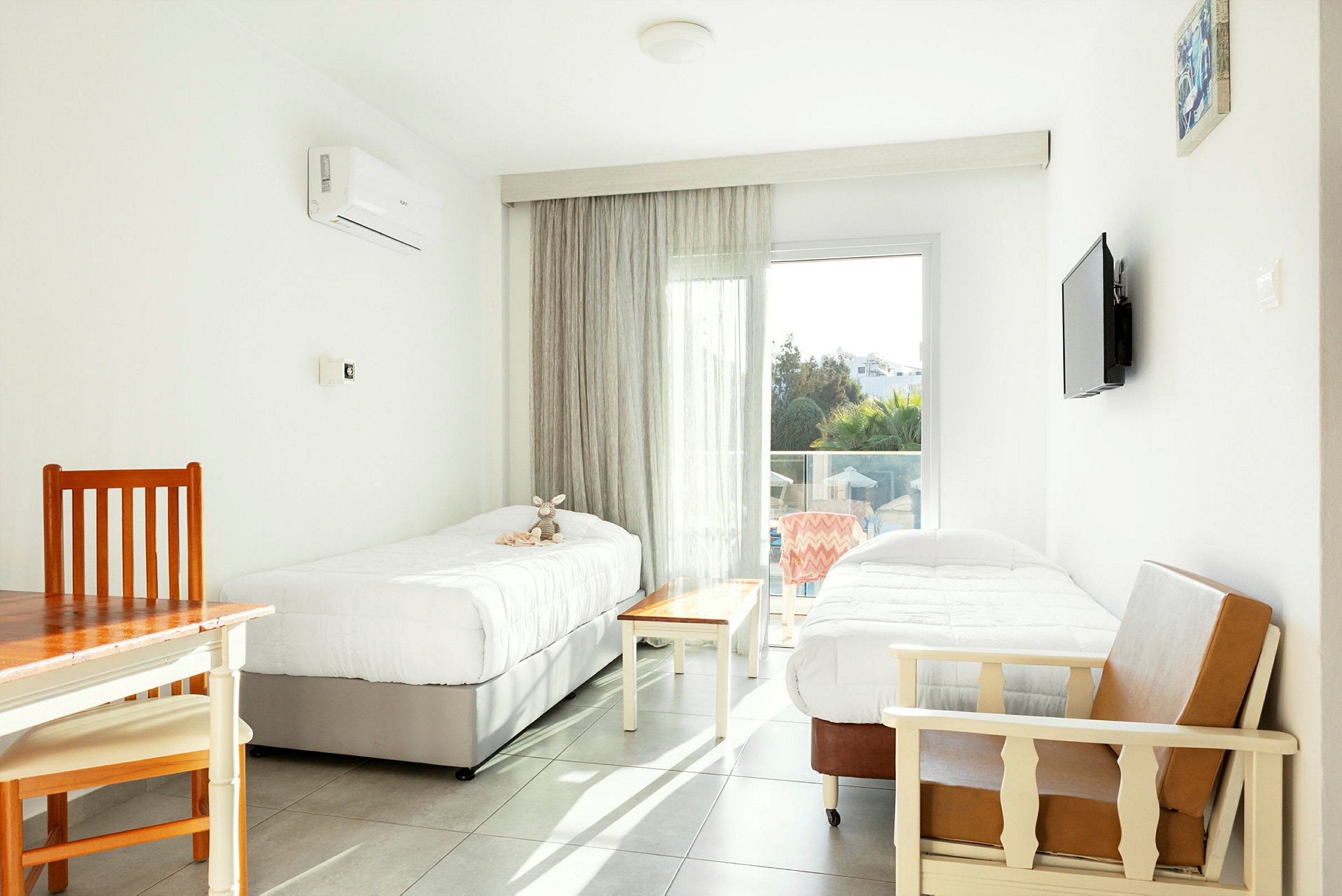 Et lyst hotelværelse med to enkeltsenge, et bord og stole samt en glasdør, der fører ud til en balkon. (AI-genereret tekst)